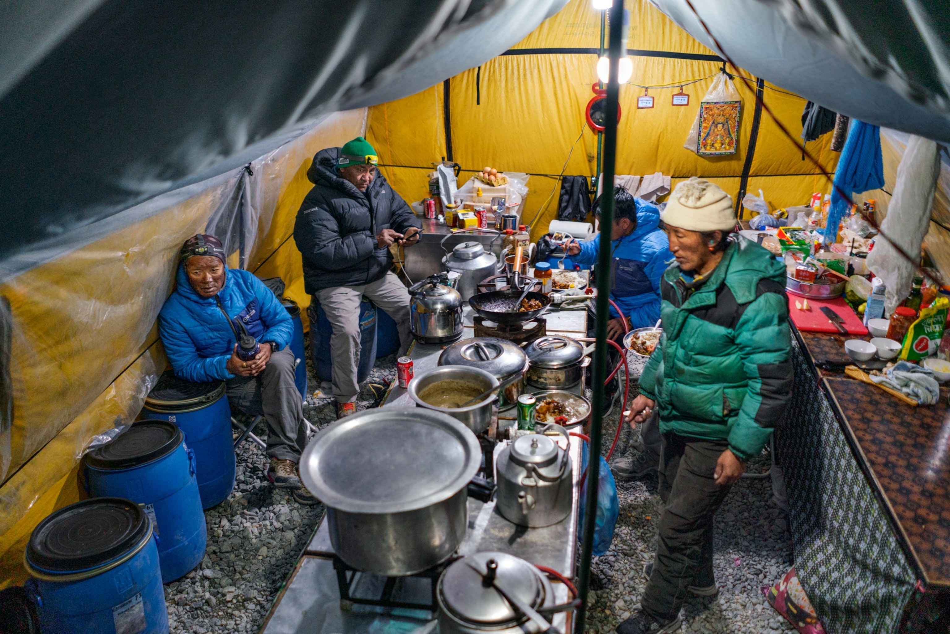 a group of people surrounding cooking materials, covered by a yellow and gray tent