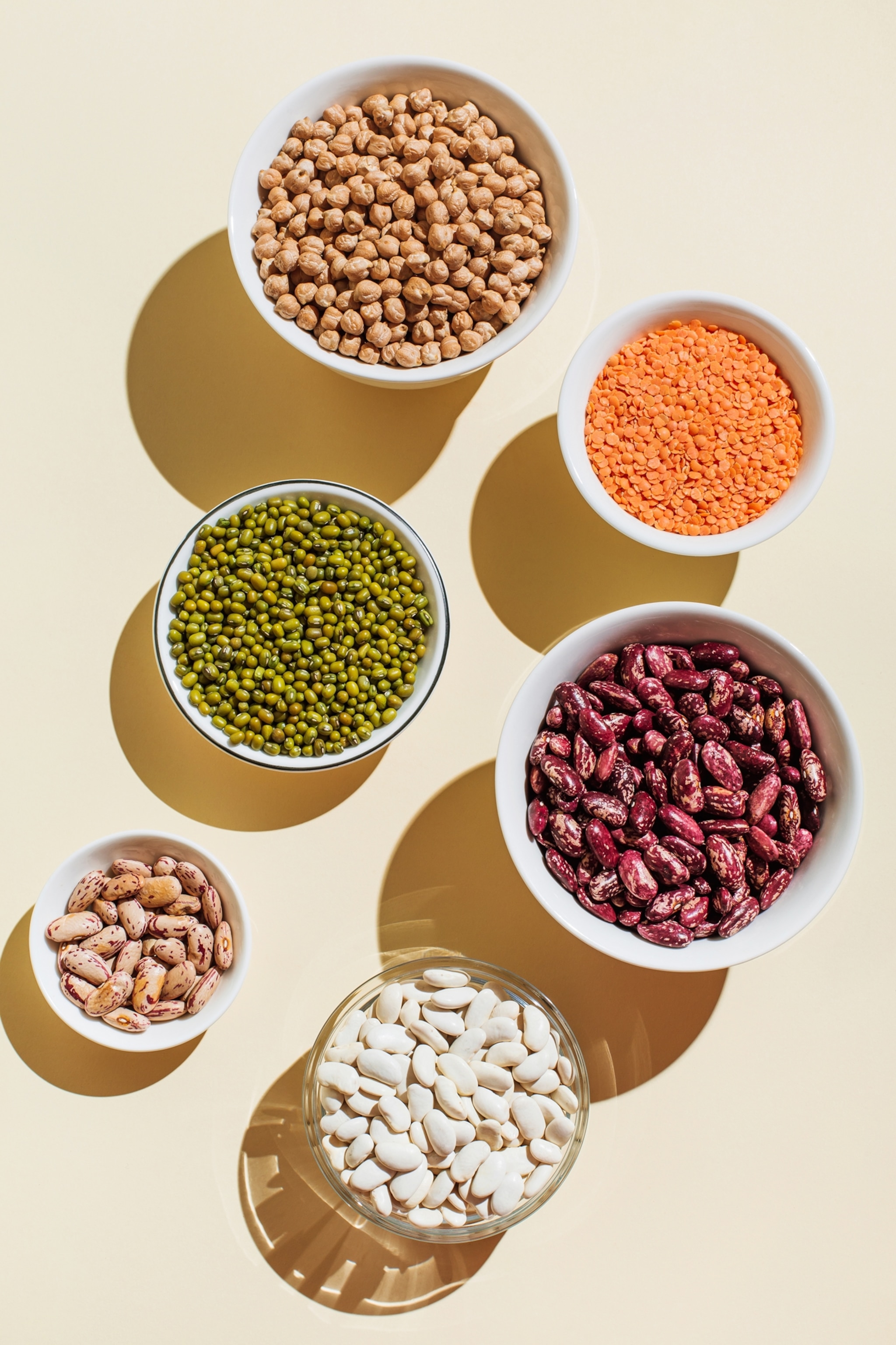 Top view of green mung beans, chick-pea, red lentil, kidney bean, almonds, and hazelnuts in bowls.