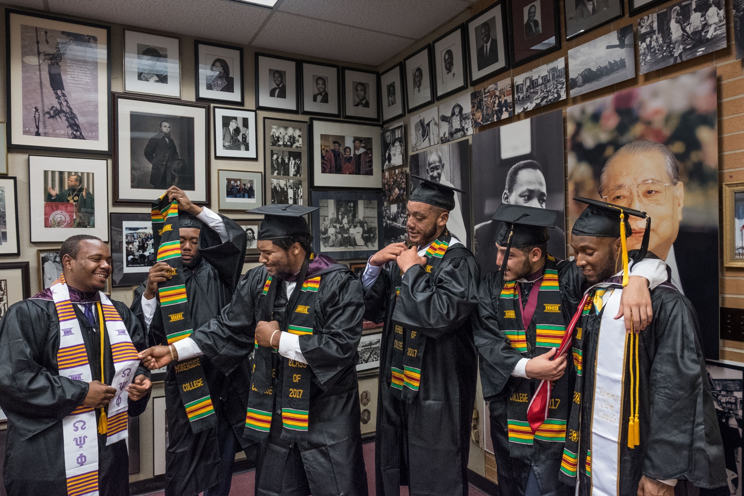 6 young black students in black graduation gowns, framed photos fill the back walls