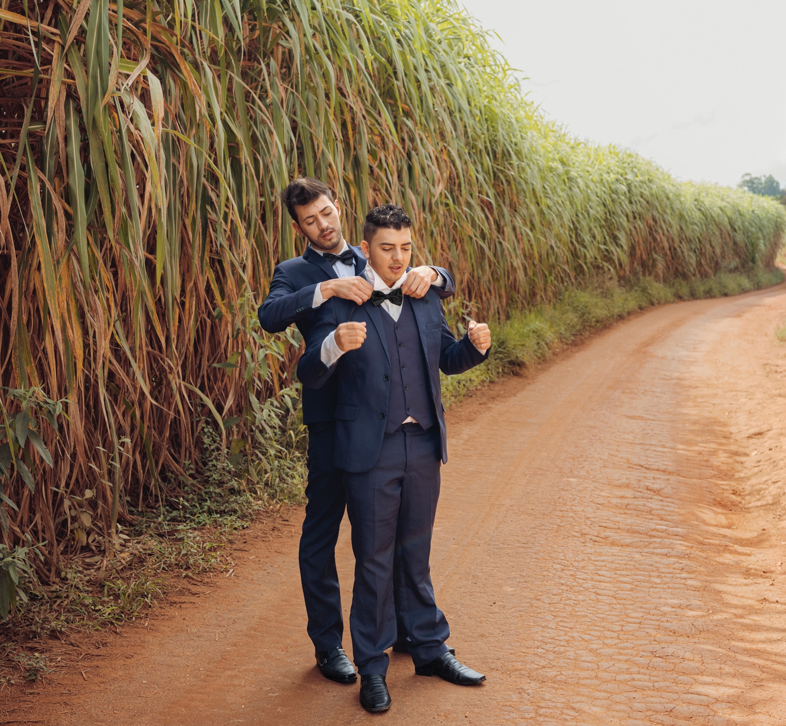 Two men stand on a dirt road next to a field of very tall plants. One man stands behind the other helping him adjust his bowtie, both men are wearing suits.