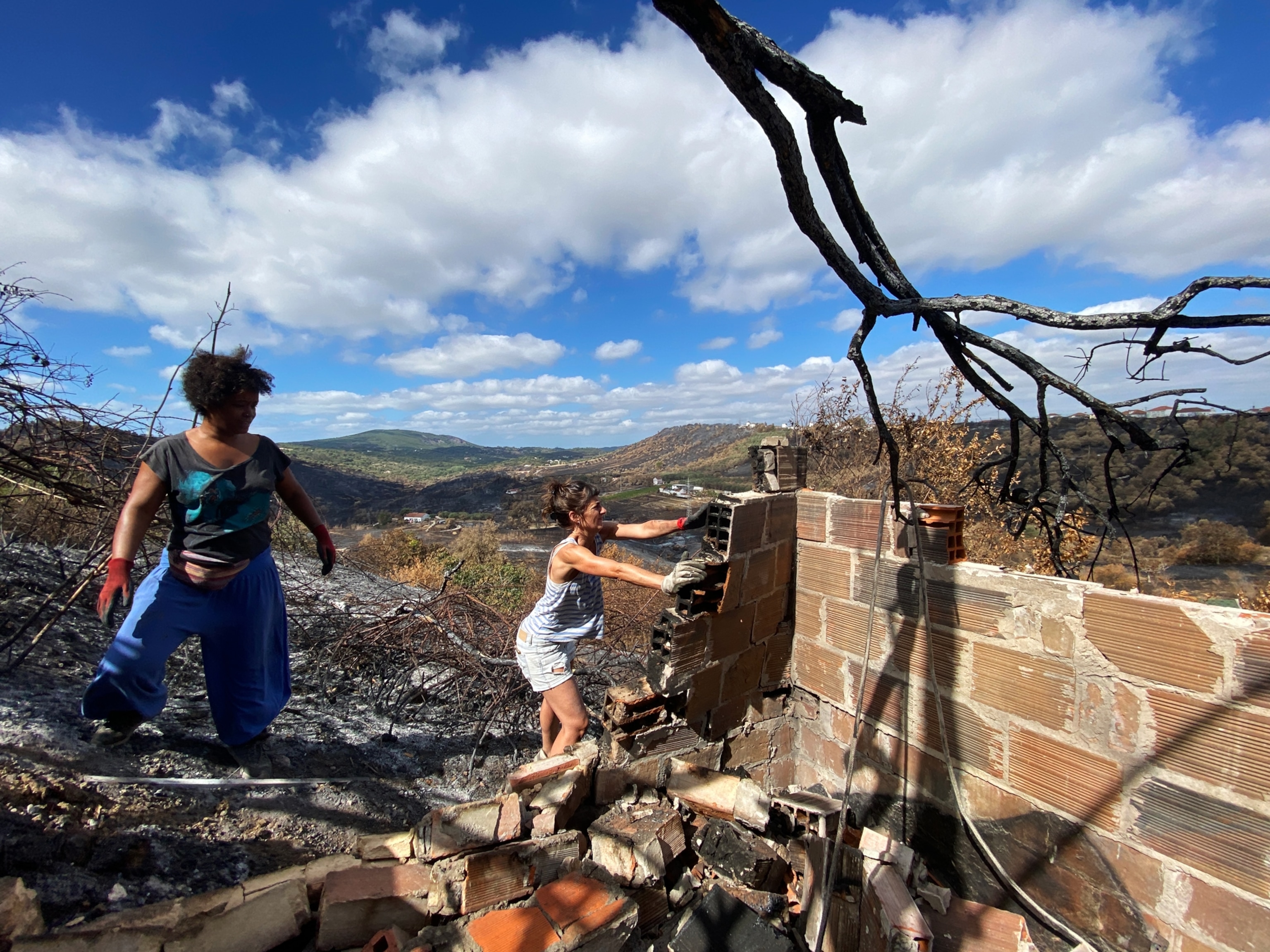 two people clean up around a falling brick wall surrounded by charred remains of the forest