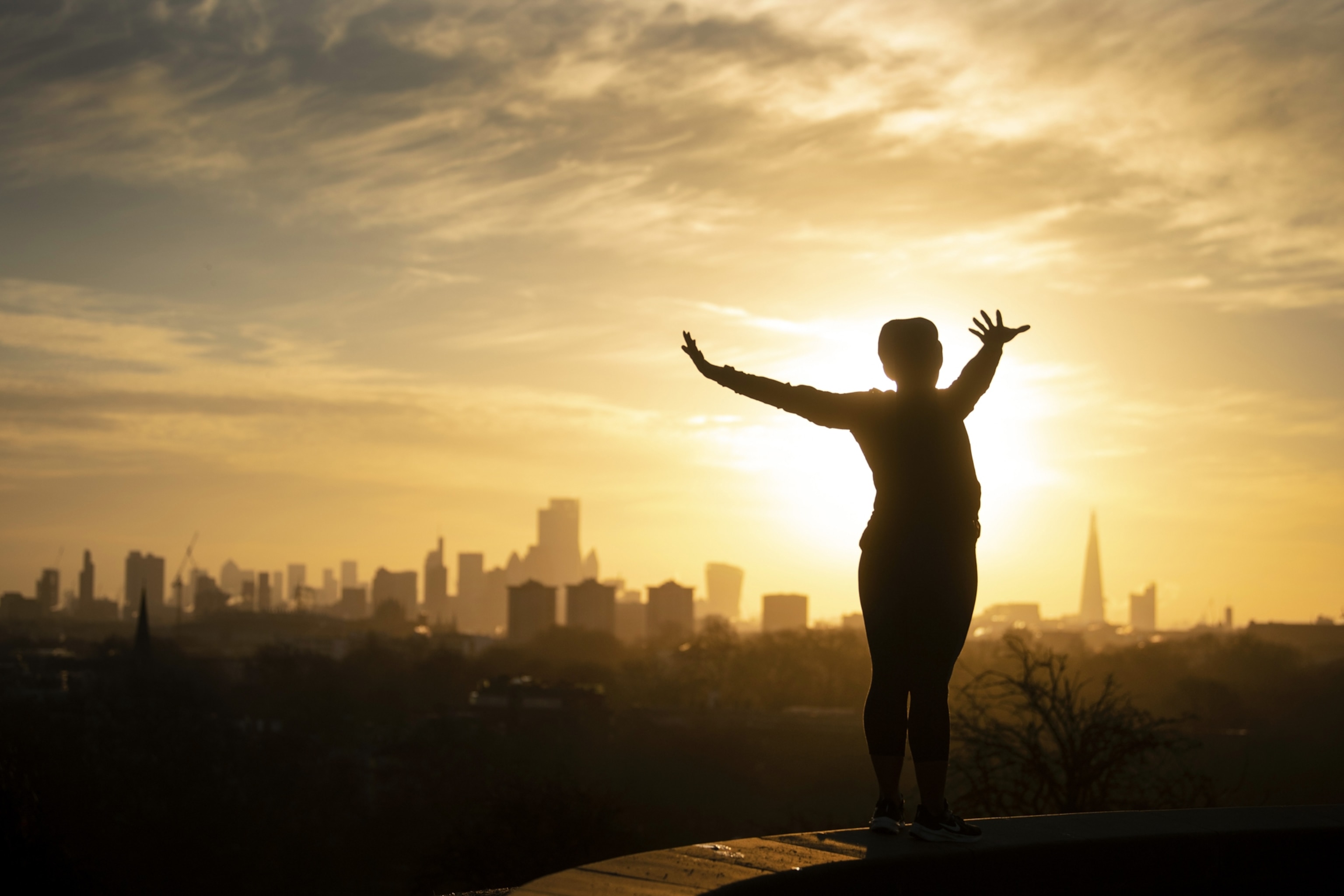 A woman is silhouetted as she performs yoga stretches on Primrose Hill, London as the sun rises over the city skyline