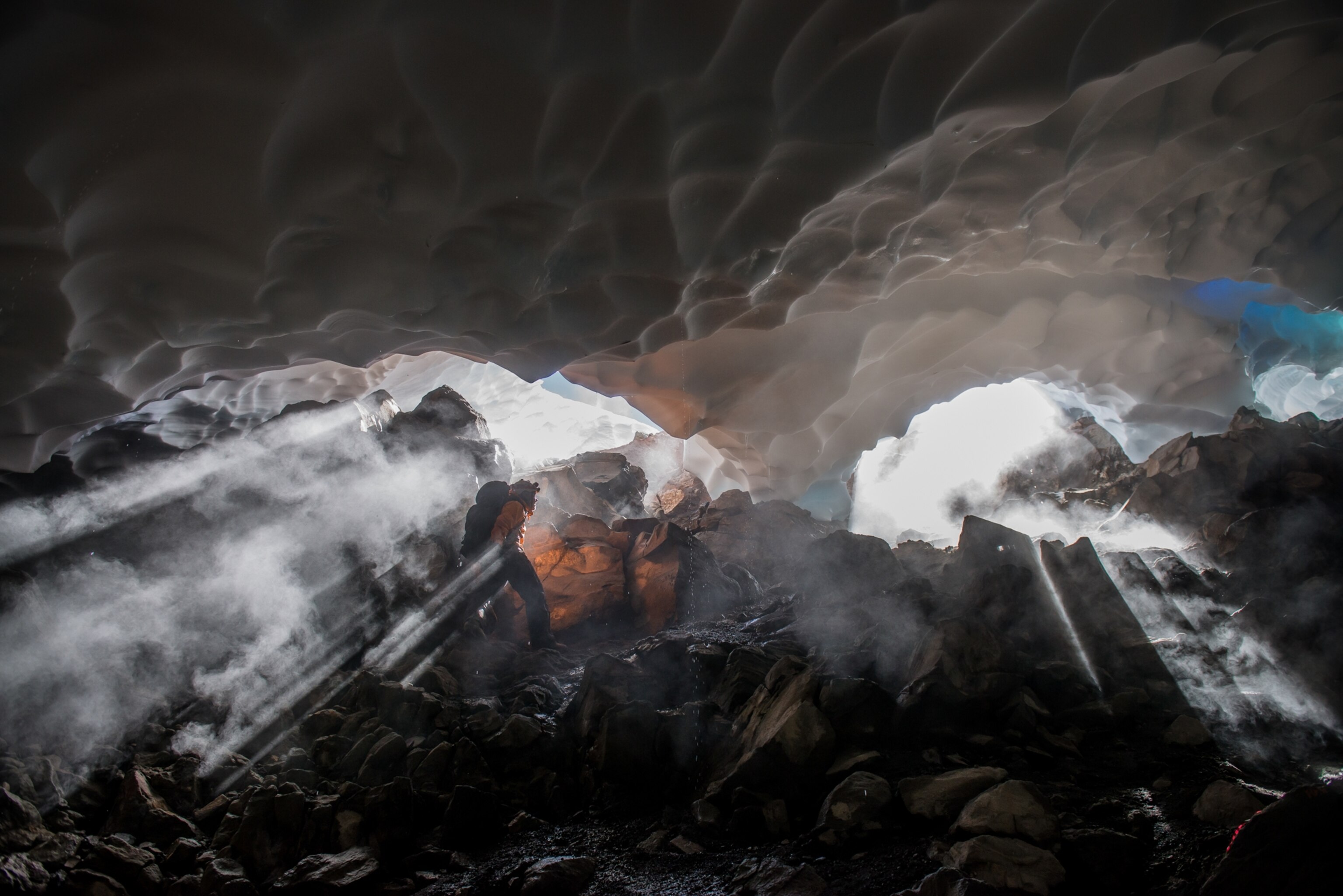 An expedition member exits one of the ice cave entrances; sun beams filter through fog created by the cold air meeting the warmer rocky floor.