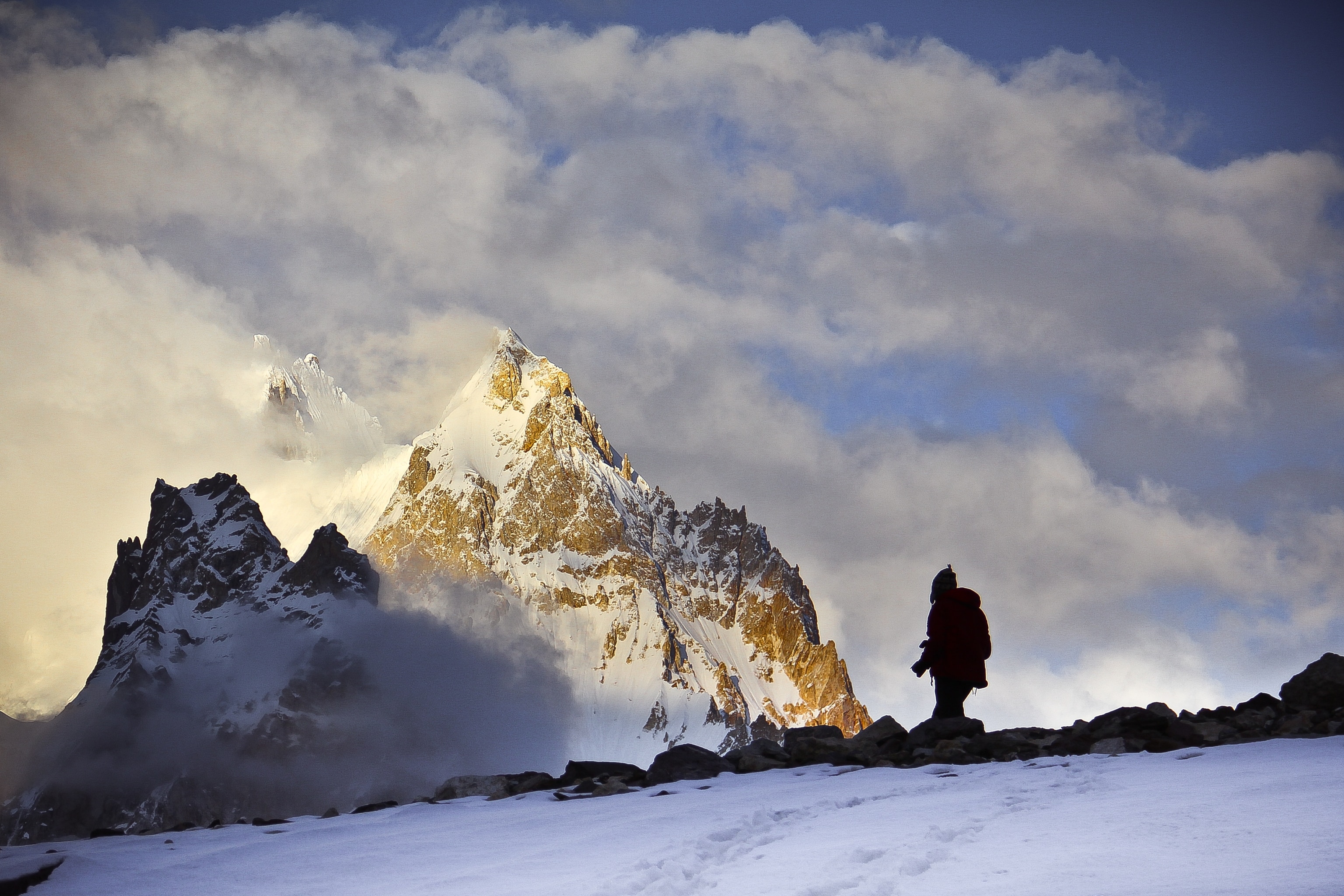A silhouetted person stands in the snowy foreground with the Gasherbrum peaks in the background