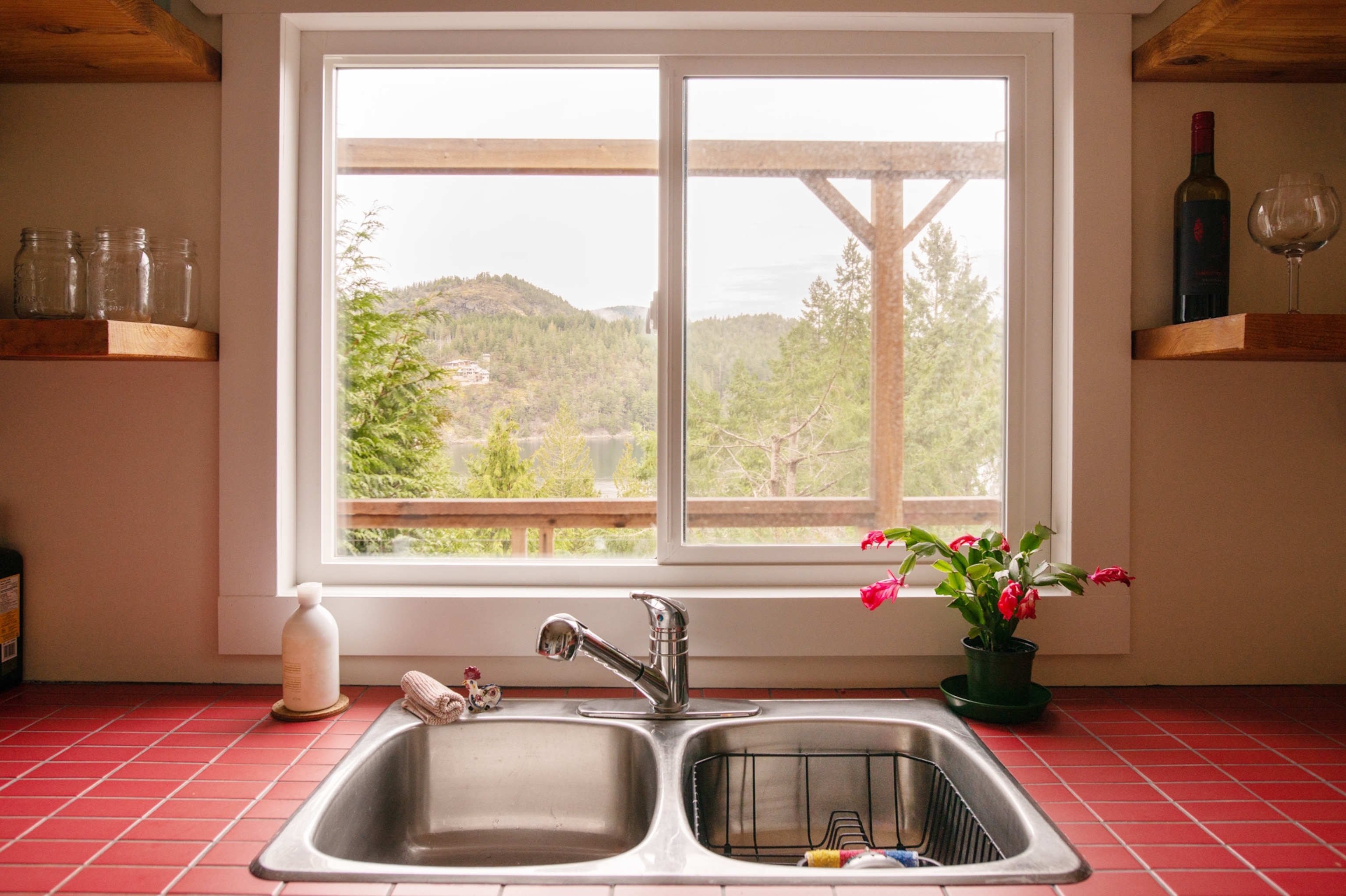 A kitchen sink beneath a window with a scenic view of tree-covered hills and a lake.