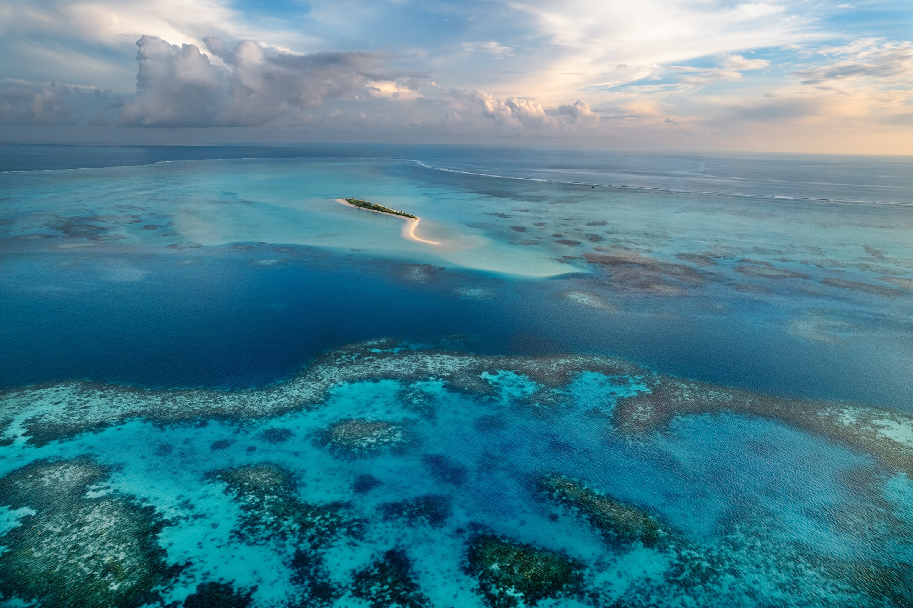Underneath a cloudy sky, a small and narrow sandbar stands out against the vibrant blue hues of the surrounding ocean.