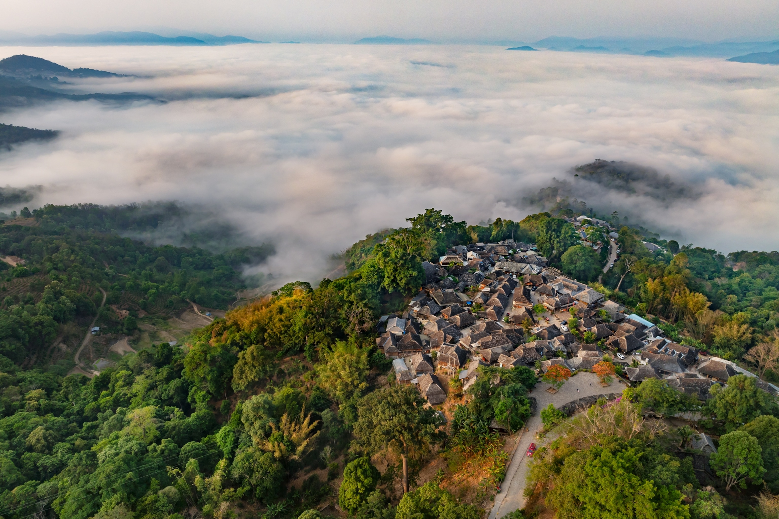 An aerial view of houses atop a mountain, surrounded by forest and clouds of fog.
