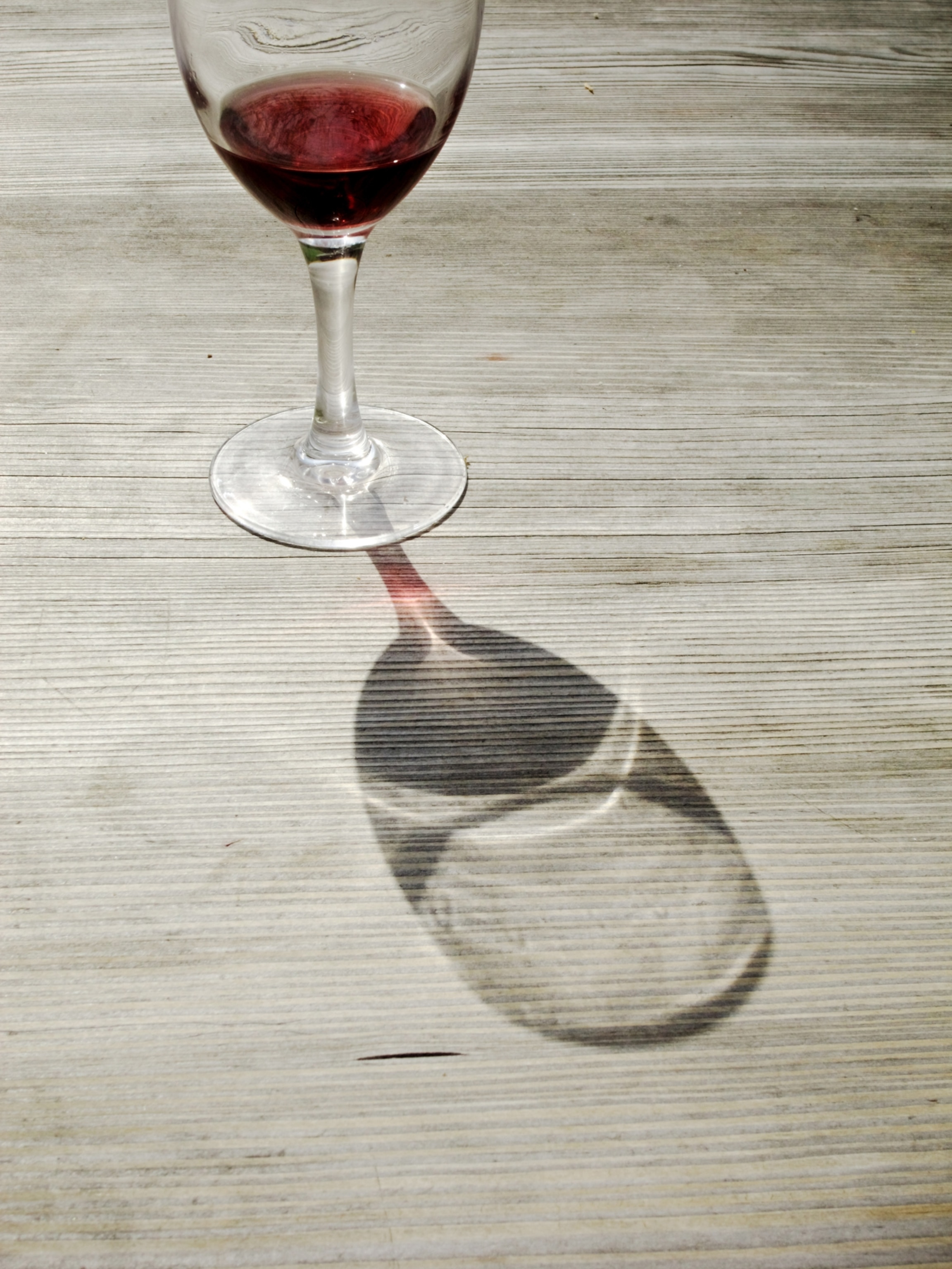 The lower half of a glass of red wine atop a light wooden table; sunlight casts a hard shadow of the glass towards the foreground.