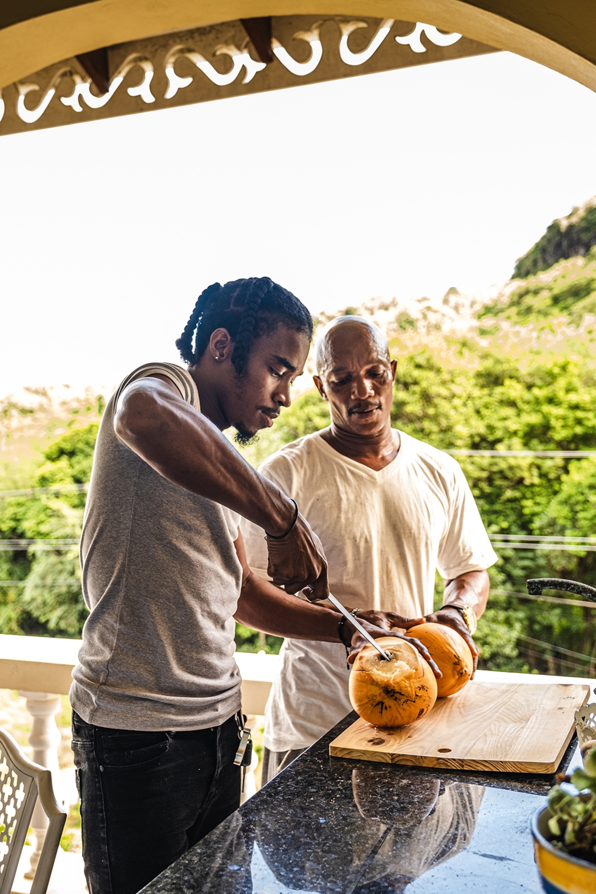 Two men carving a fresh coconut with a knife.