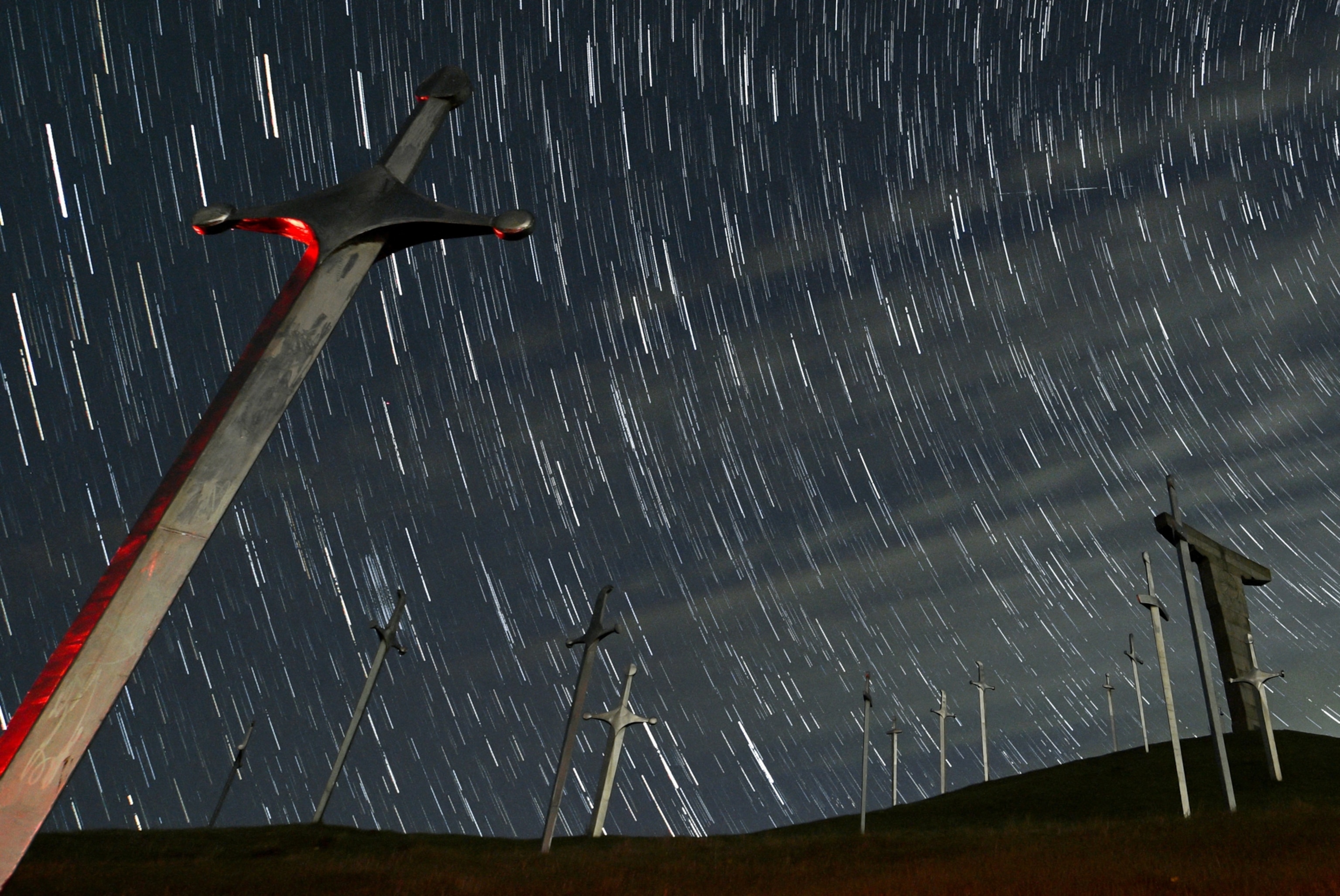 A swirl of stars trailing across the night sky, seen above a memorial monument of several large swords sticking from the ground.