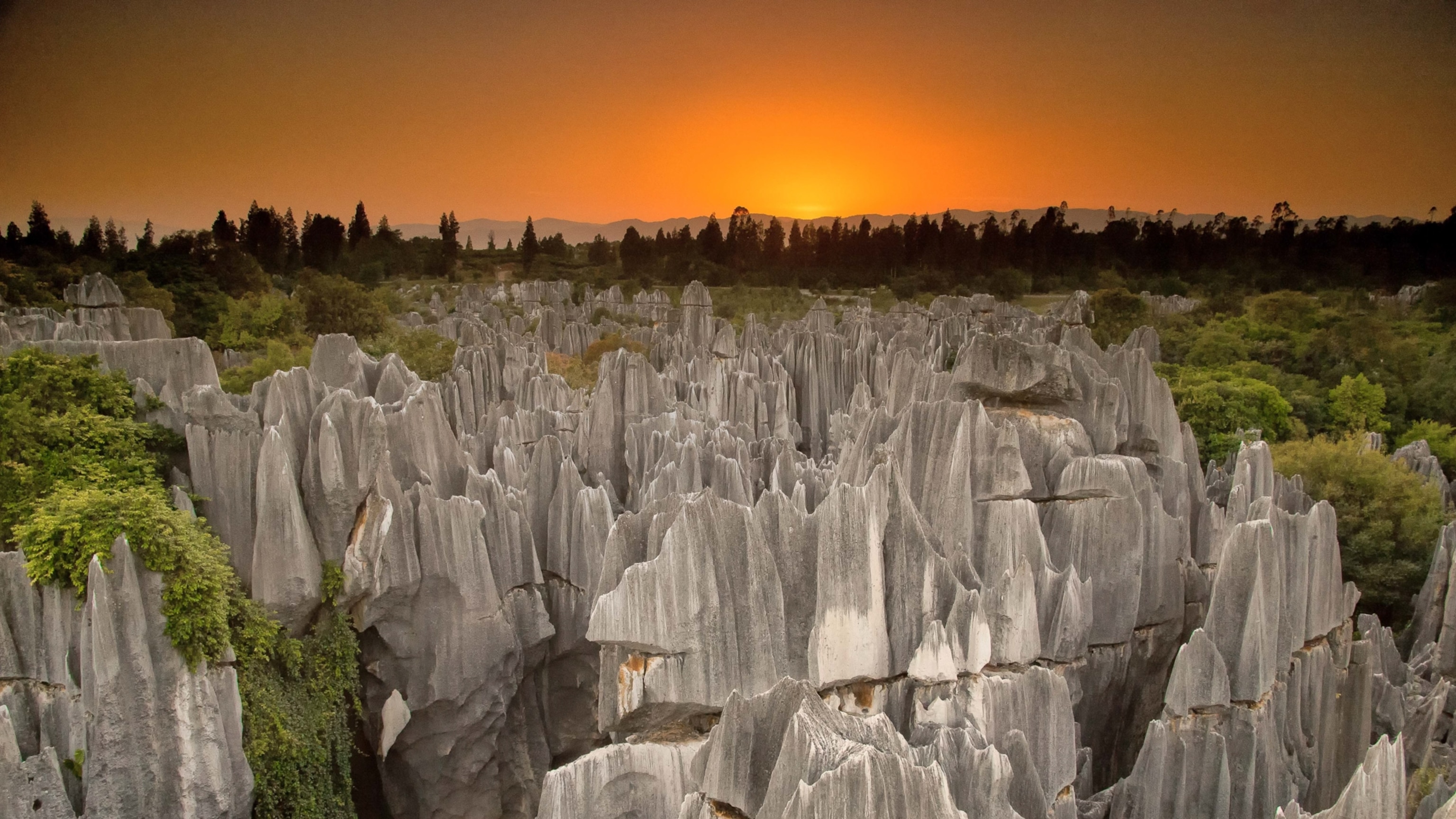 An aerial view of jagged Limestone karst formations as the sun sets on the horizon, casting an orange glow across the sky.