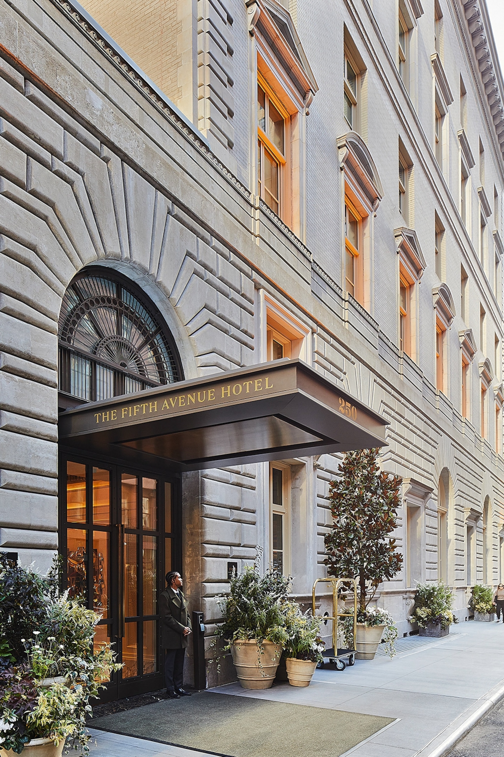 Exterior of a grand stone building with an overhang reading The Fifth Avenue Hotel with an entrance of a green carpet