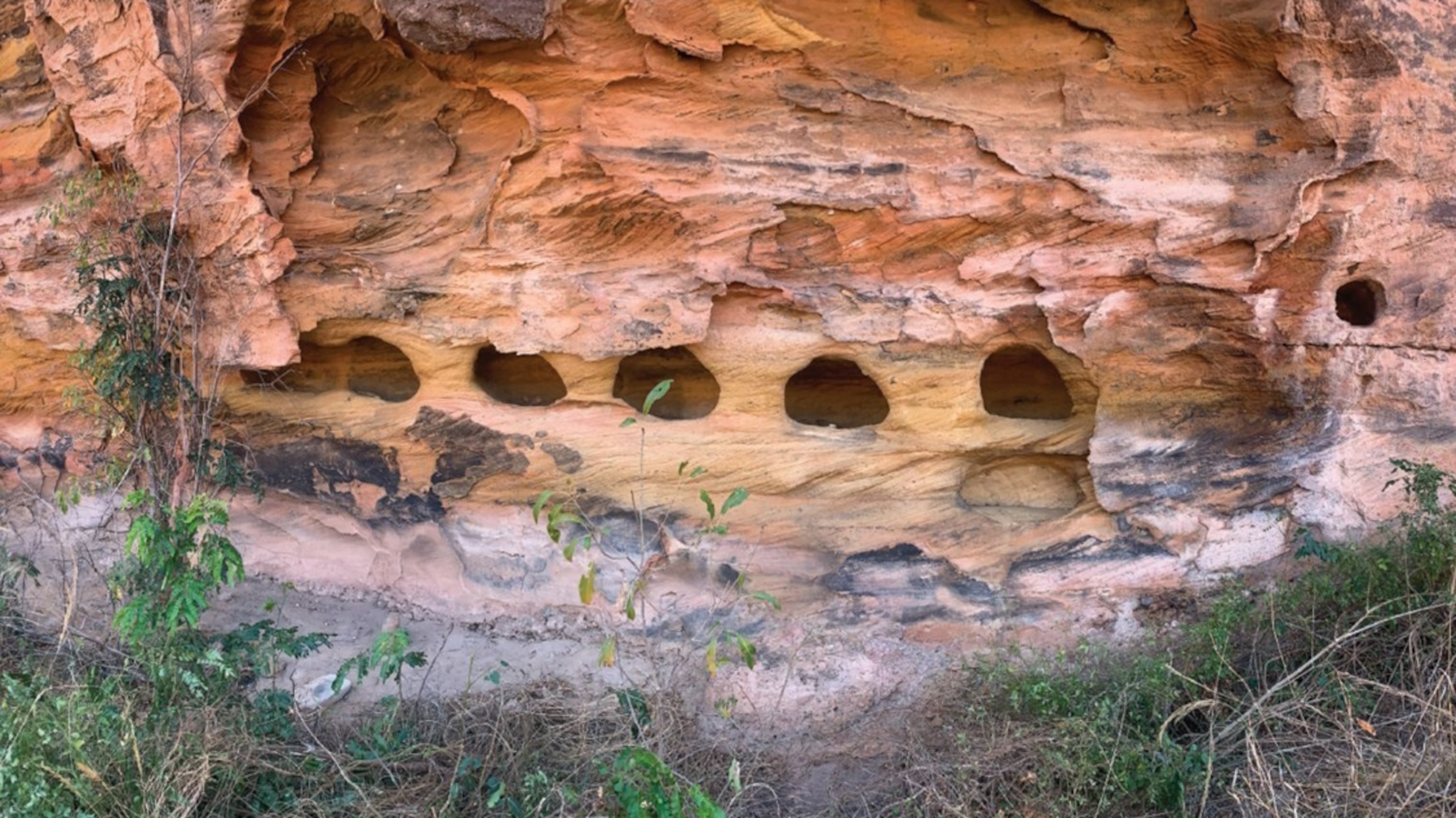 The face of a sandstone cliff, its jagged rocky surface shows hues of burnt reds, orange-browns, and pale yellow. Into the sandstone, a parallel series of 6 windows have been carved, leaving pillars of sandstone between. A smaller round window is carved into another section of sandstone, slightly to the right.