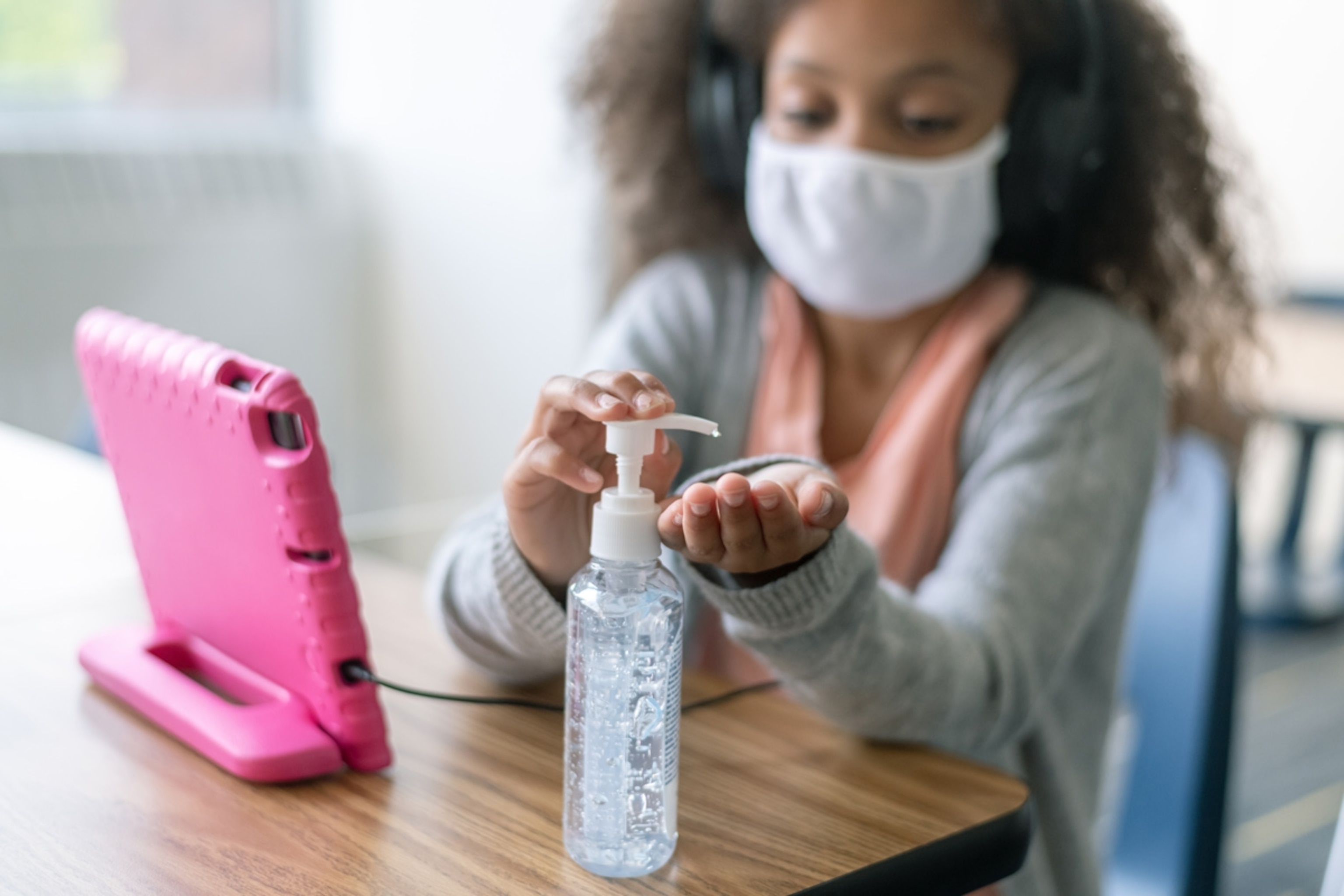 An elementary age girl of African descent wearing a protective face mask and sitting at a desk divided by a plexiglass protective barrier takes a break from her school work to use hand sanitizer.