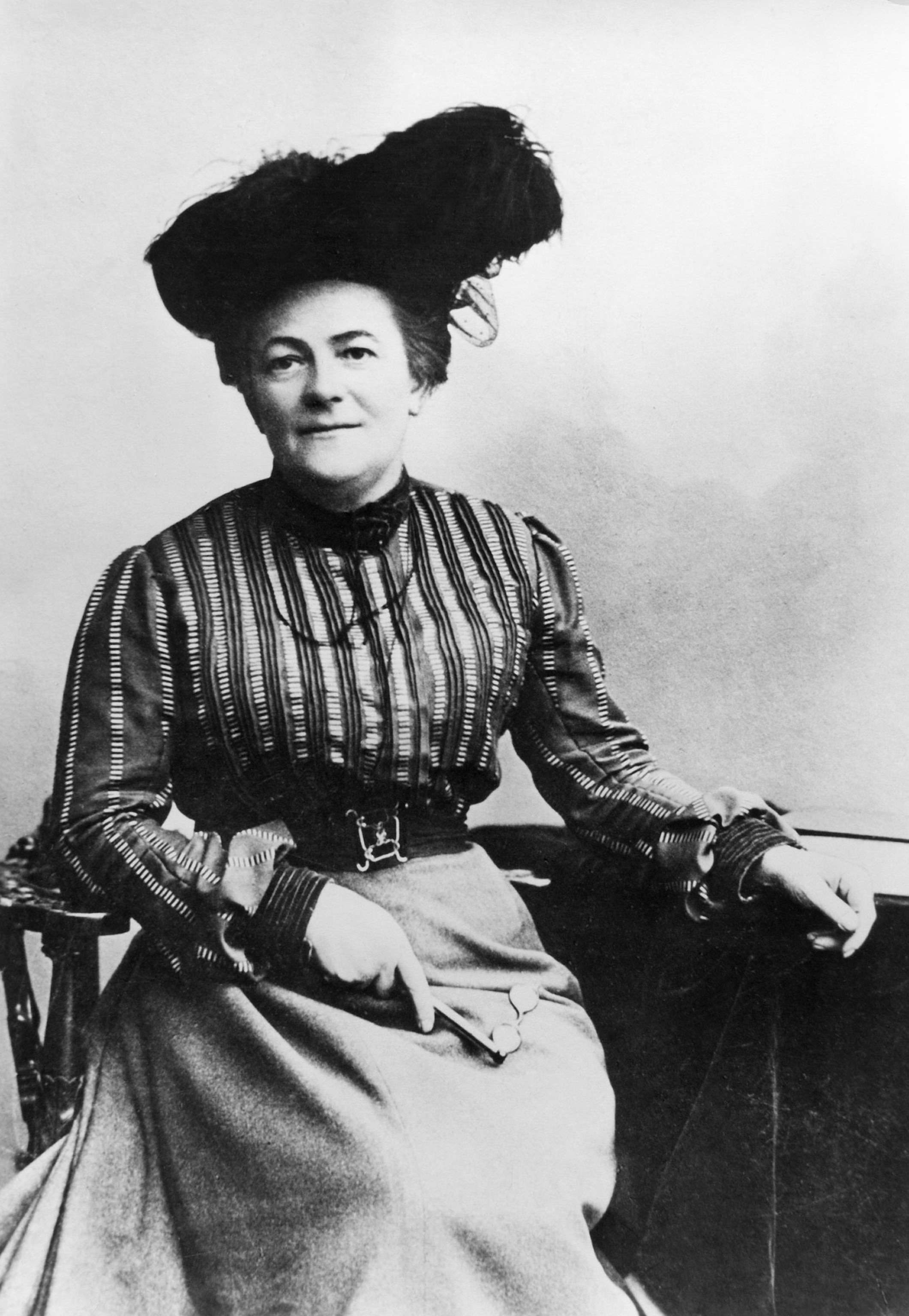 A black and white portrait of Clara Zetkin sitting on a chair wearing a large hat