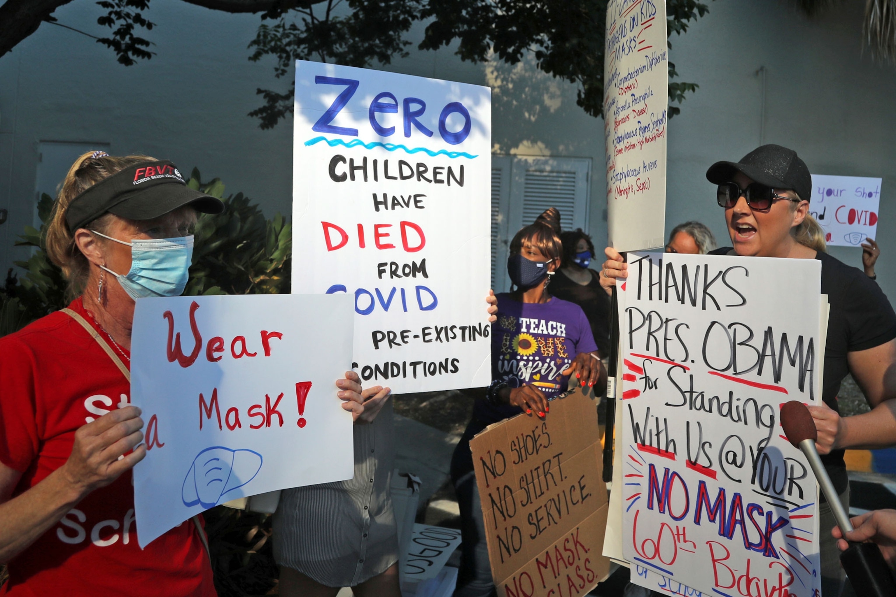 A masked woman holds a sign that reads "Wear a Mask!" and another woman faces her, holding a mask that says "THANKS PRES. OBAMA for Standing With US @ YOUR NO MASK60th Bday Party."