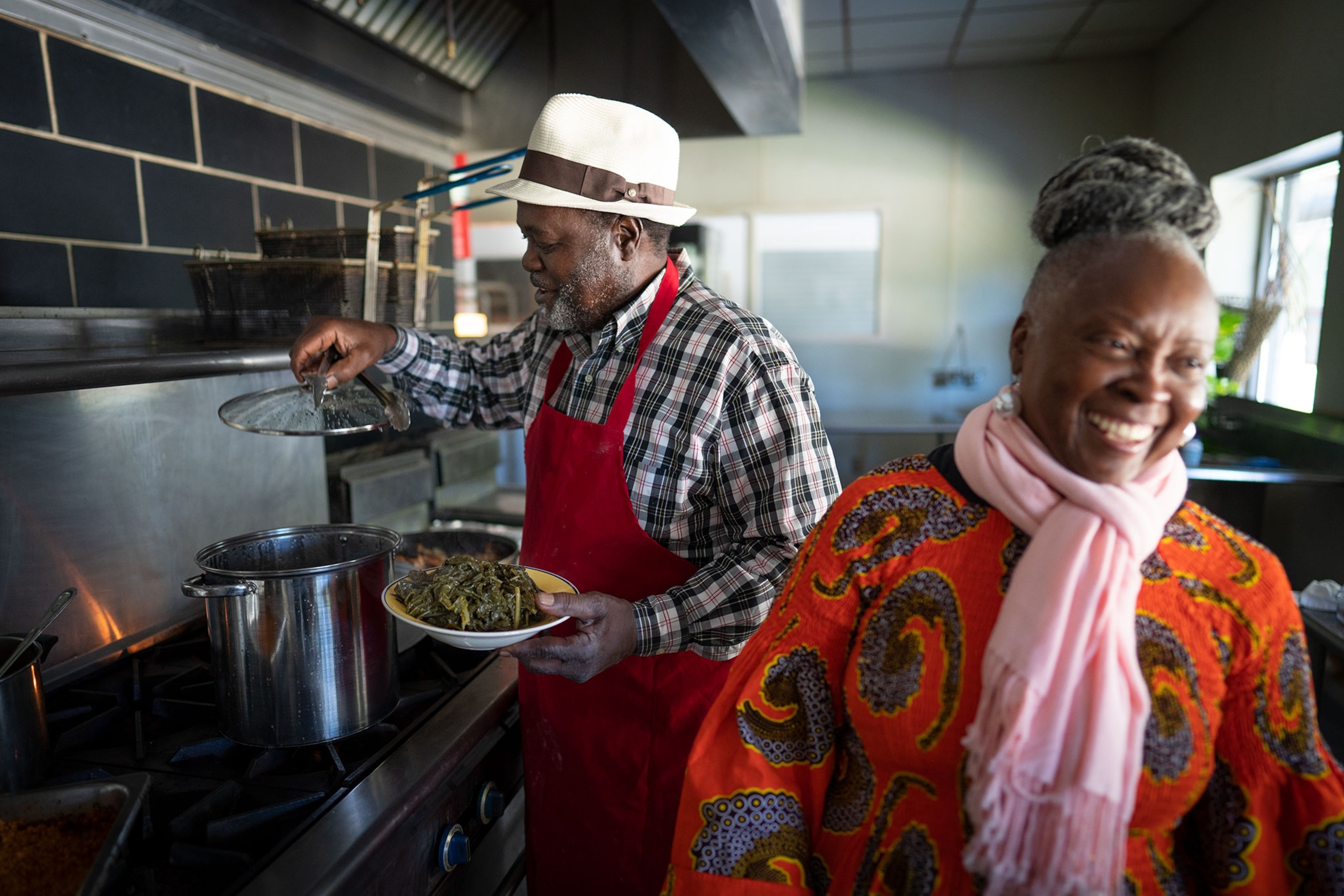 The Chef stands in front of the stove with a plaid shirt and red apron, with a pot lid in one hand and a dish pilled high with greens, his wife stands near with an orange shirt and pink scarf smiing gleefully.