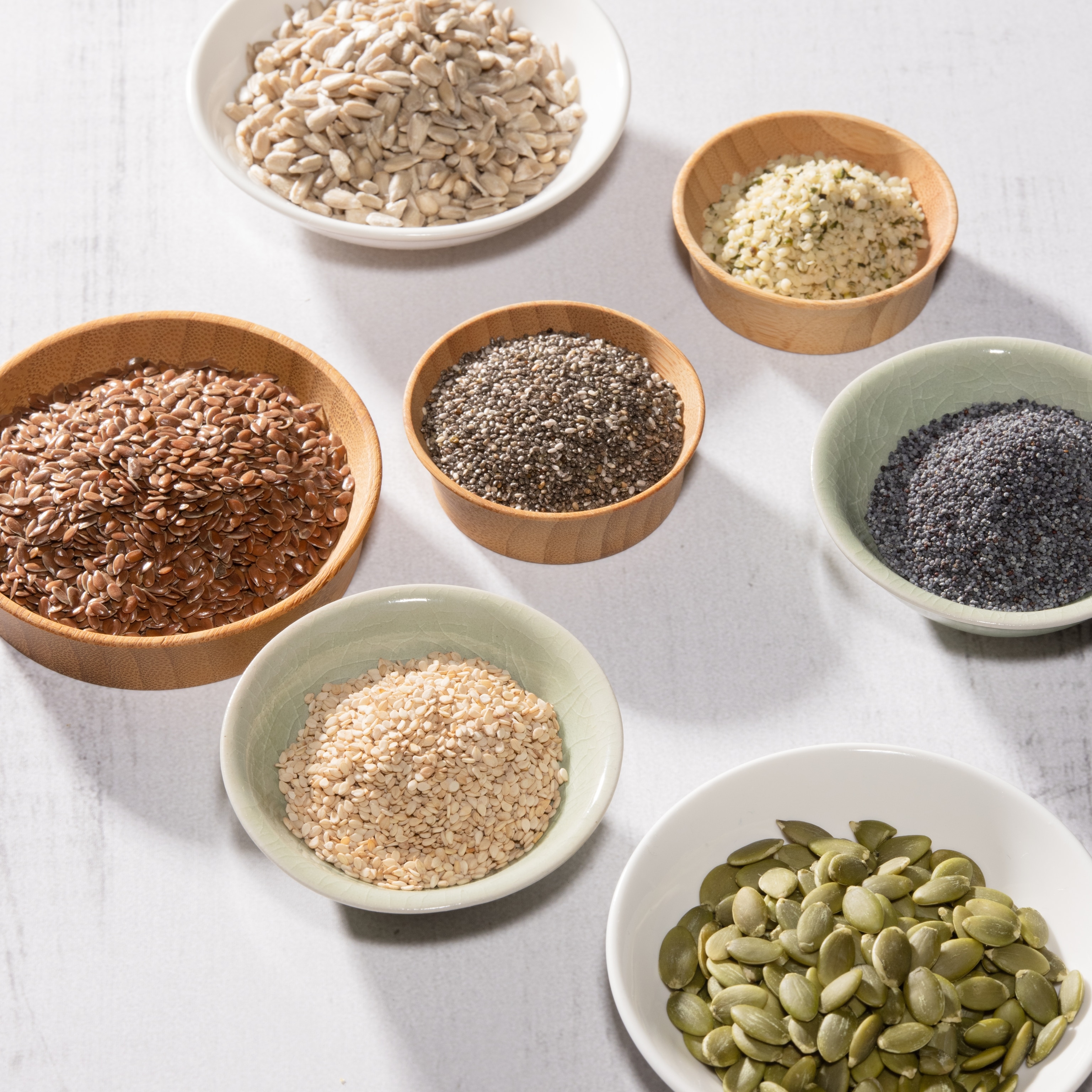 Seeds are displayed in various bowls atop a white wood background.