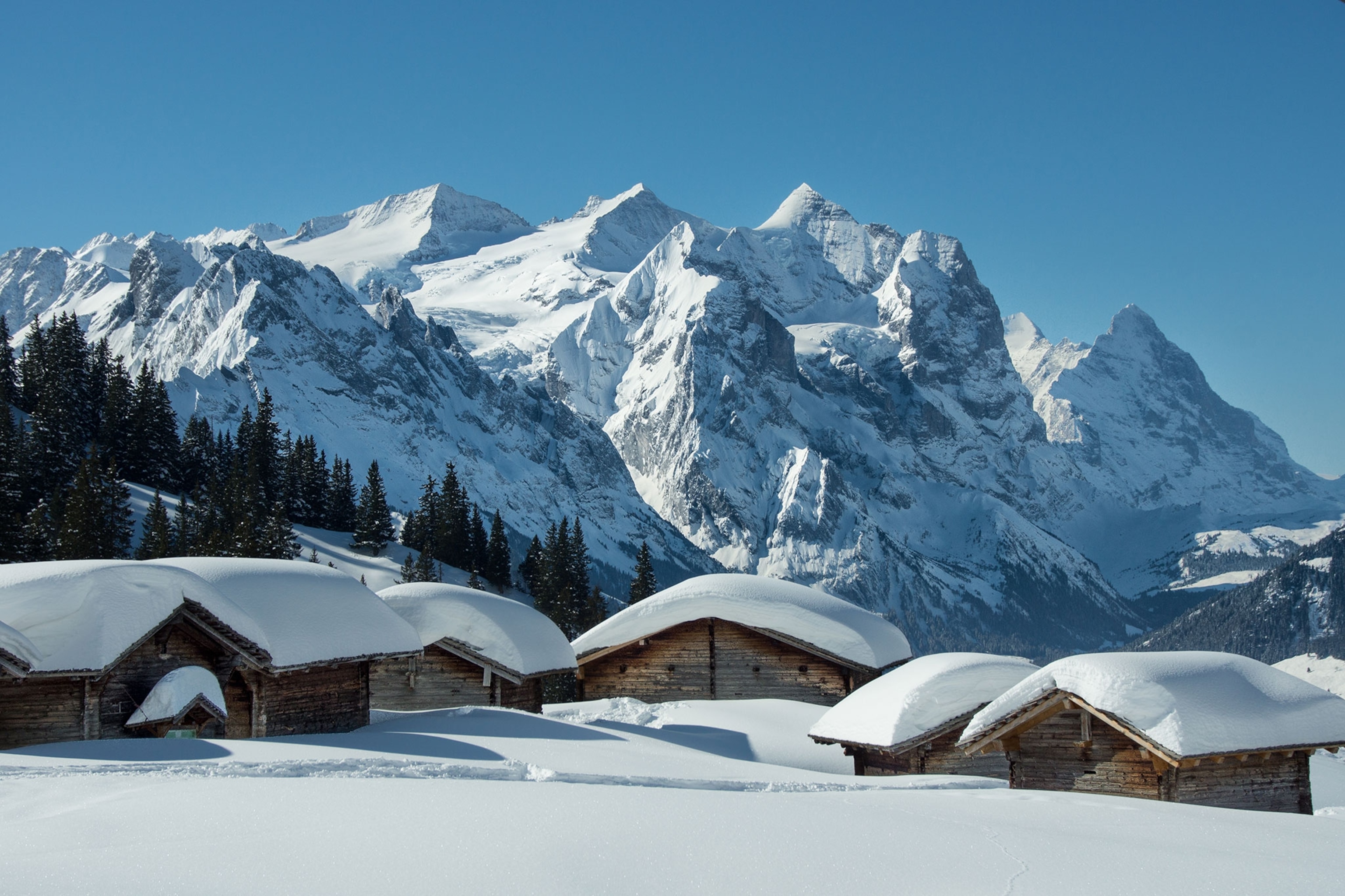 Cabins with snow covering their roofs sit in front of snow-capped mountain peaks.