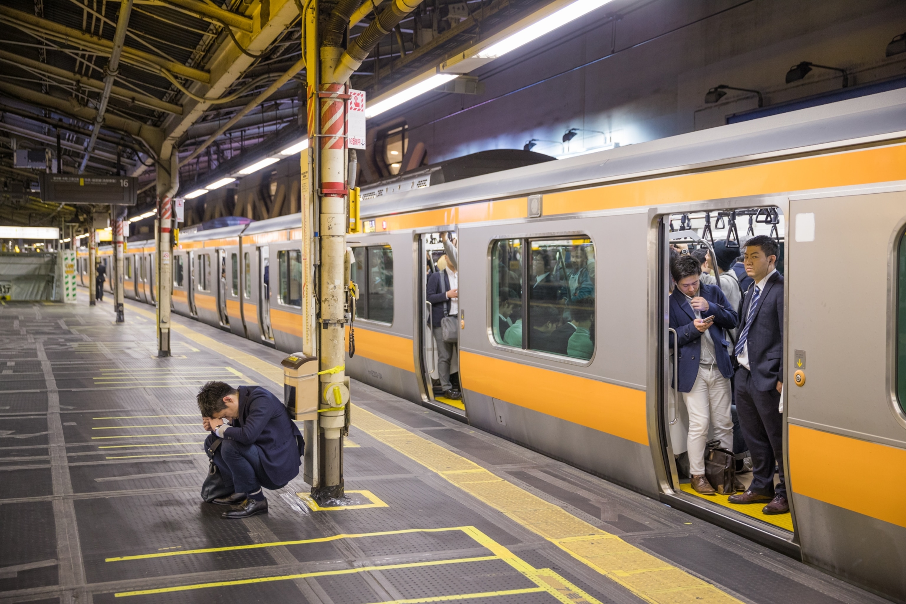 a man squatting down a resting on a pillar in the subway as a train waits to depart
