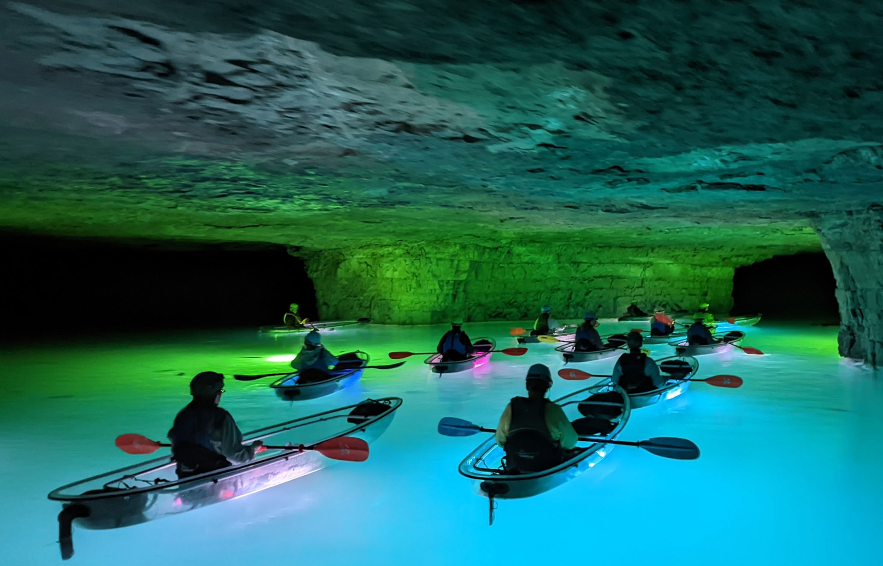 Kayakers underground paddle towards a smaller cave opening on glowing blue waters.