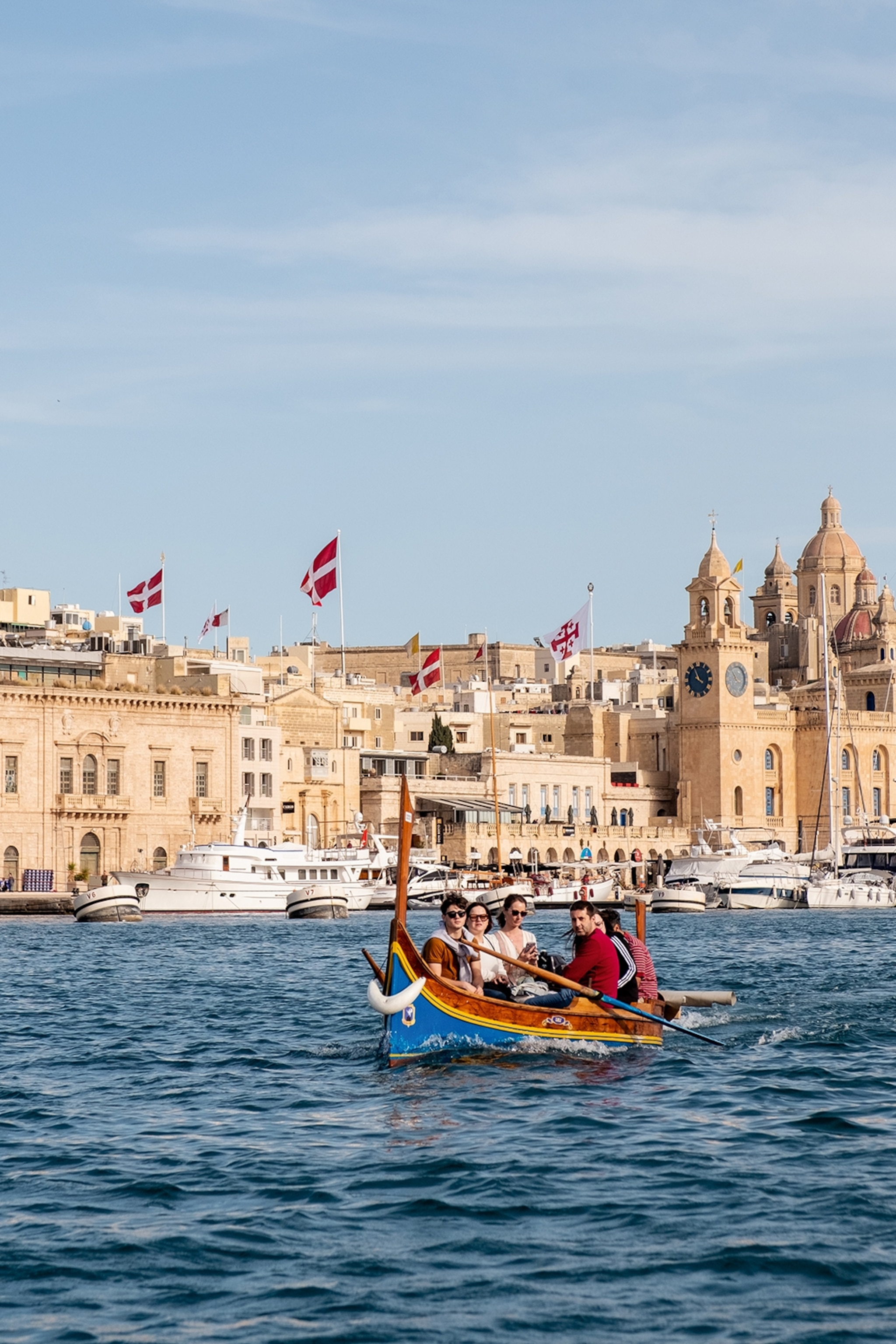 A group of people exploring Valletta in Malta by a traditional wooden boat. The sea is dark blue and choppy, whils in the background bigger yachts are docked in front of honey-coloured buildings.