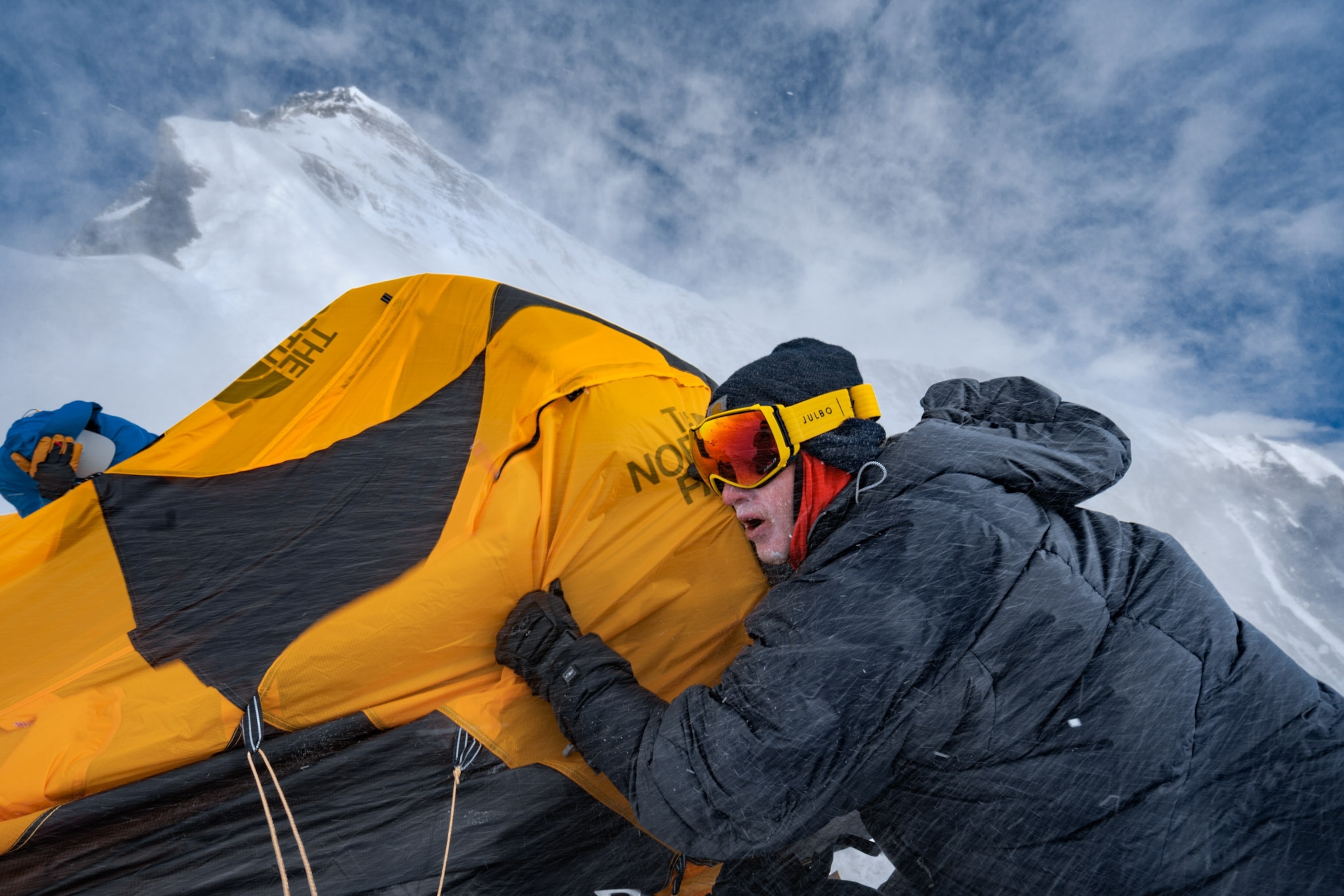 a person in ski goggles clinging to a tent as snow dusts around him