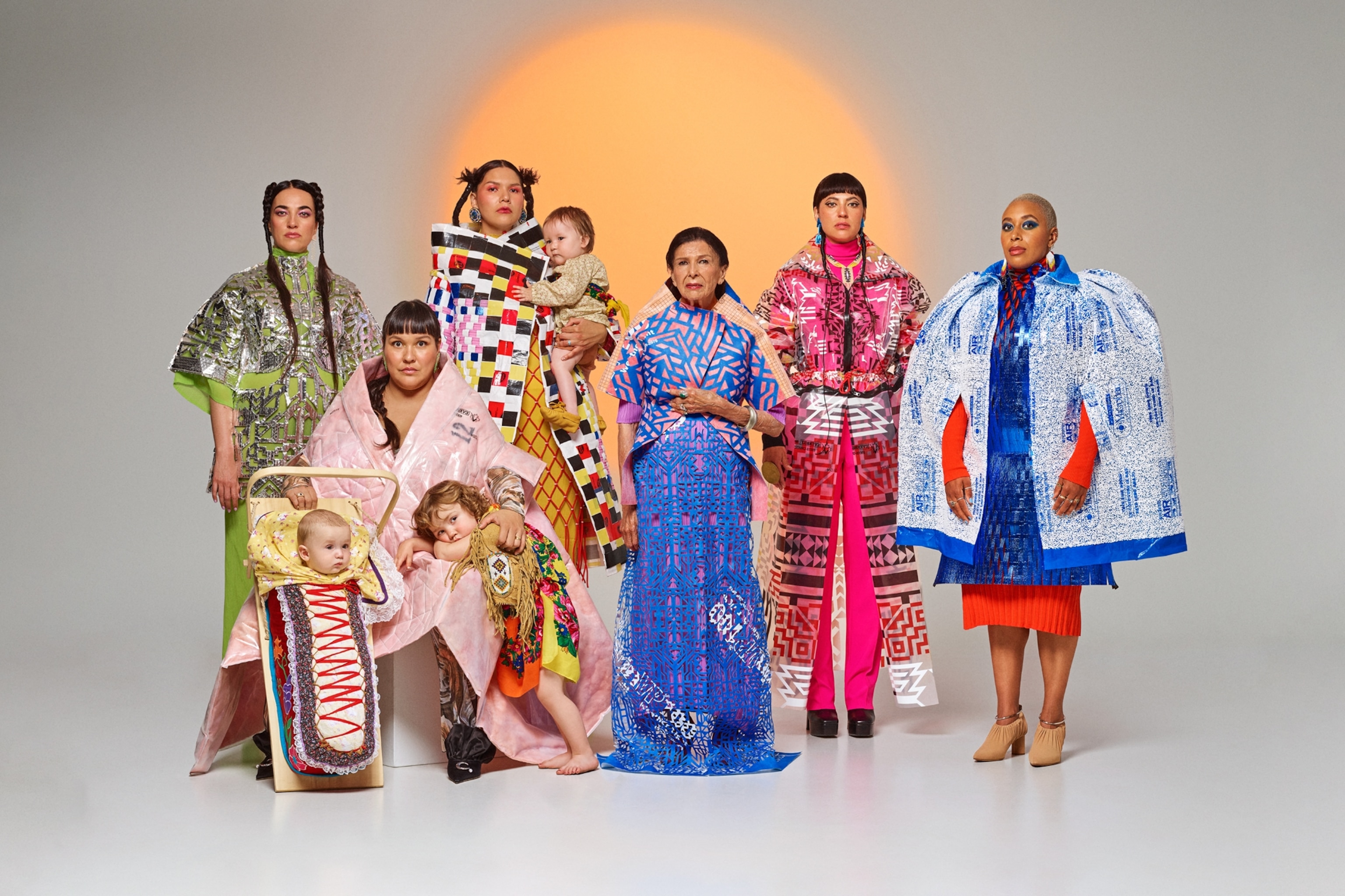 Six women and two children dressed in futuristic but yet traditional costumes posing again light gray background with large golden circle representing the sun.