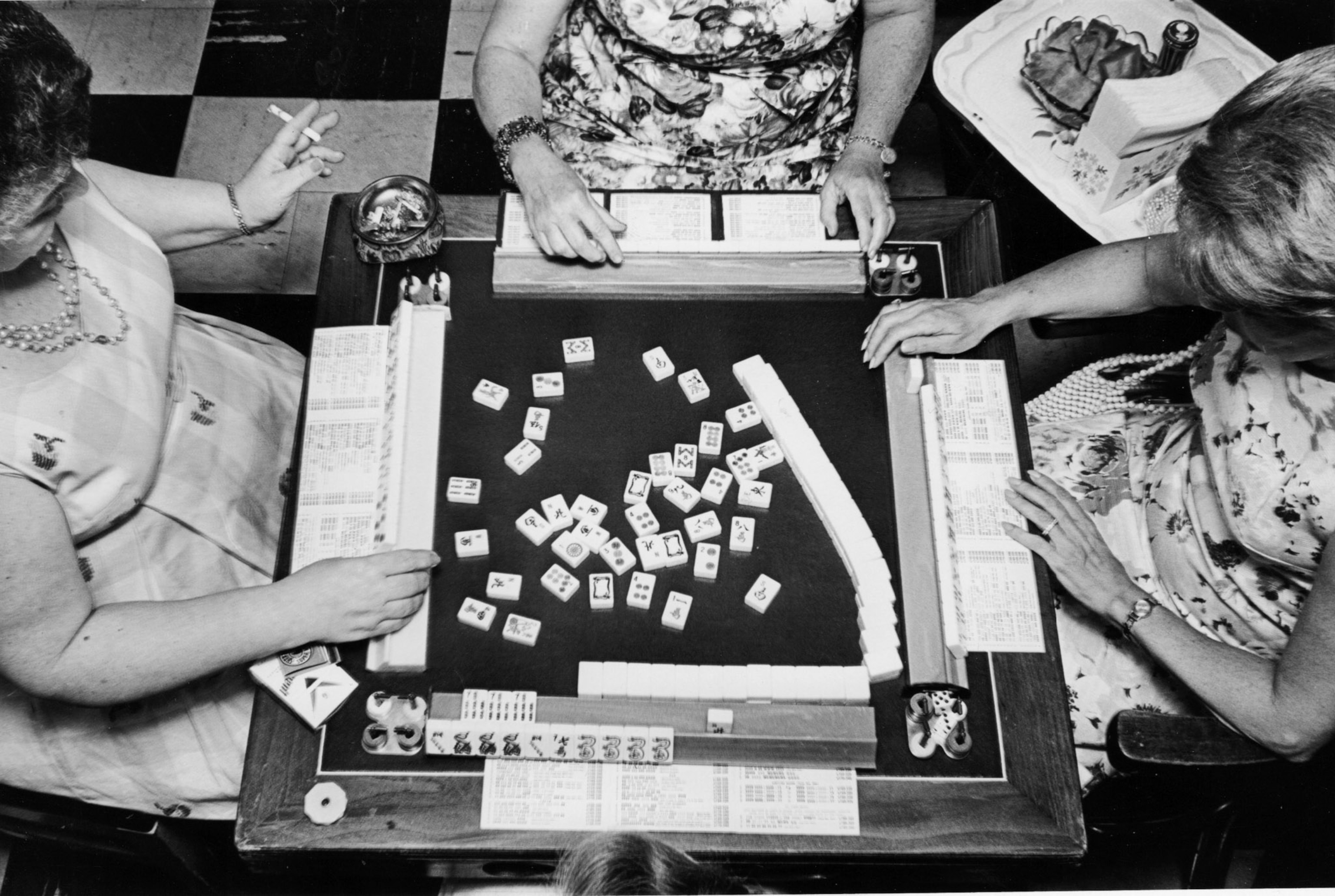 A black and white image of an aerial view of a mahjong table with women playing the game drinking and smoking.
