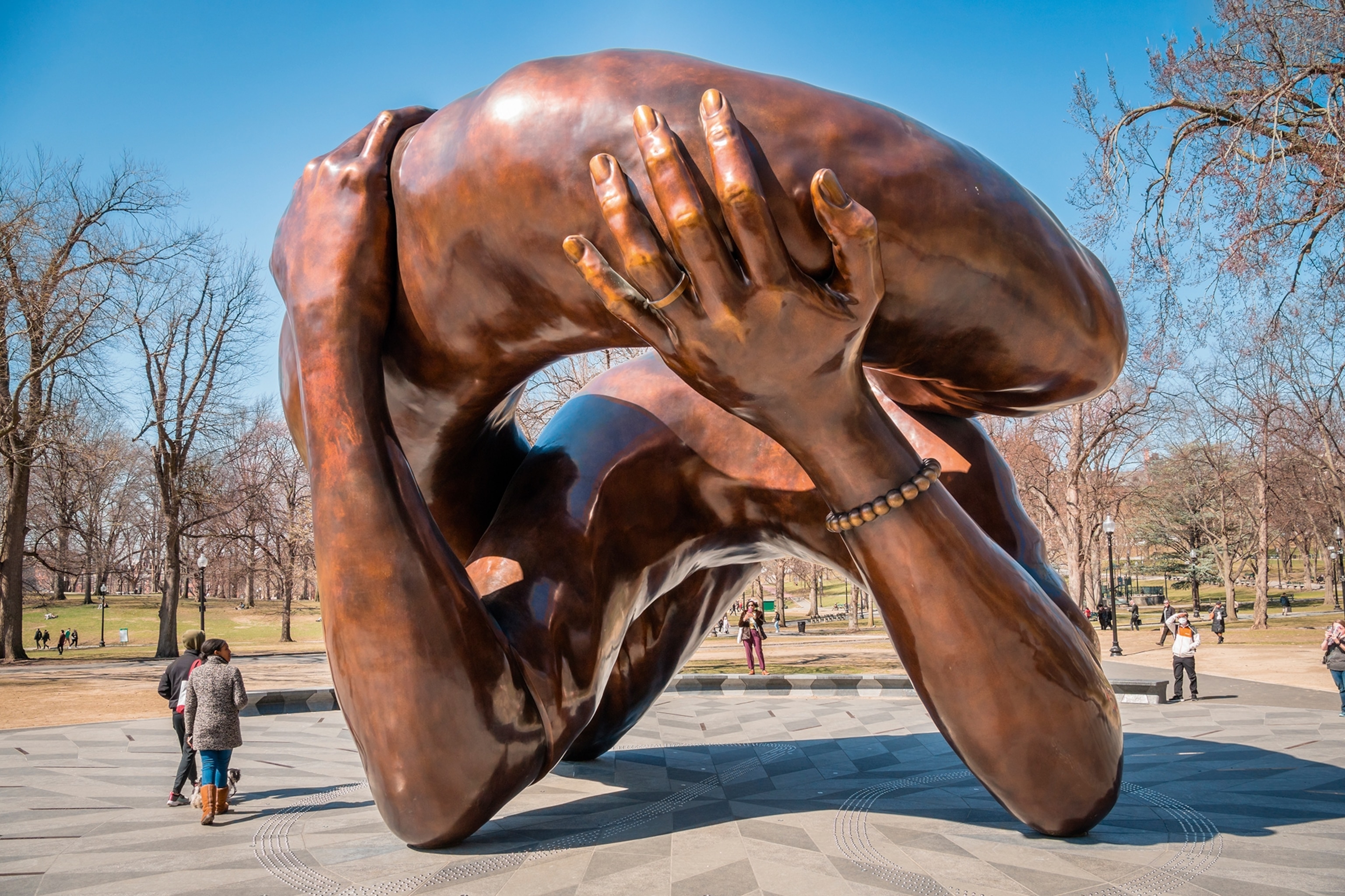 An imposing, abstract brass sculpture in a park, reminiscent of connected yet hugging arms.