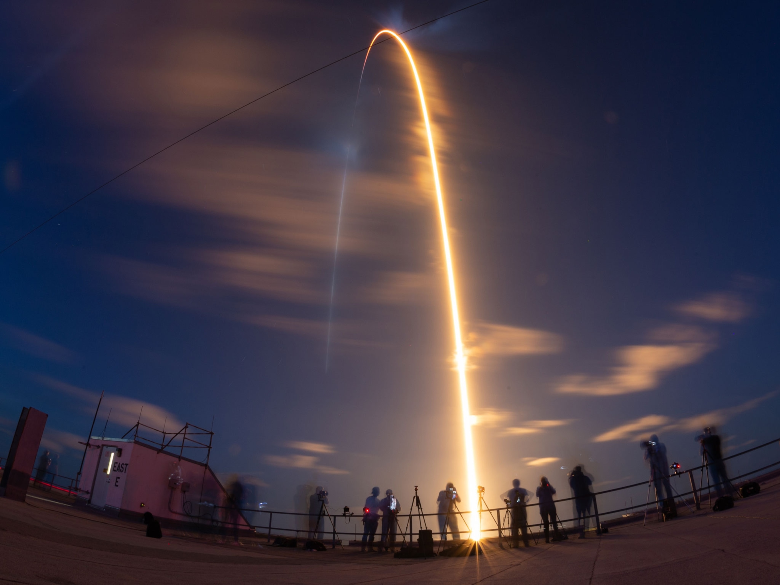 A SpaceX Falcon 9 light stream arcs in the sky above a group of photographers.