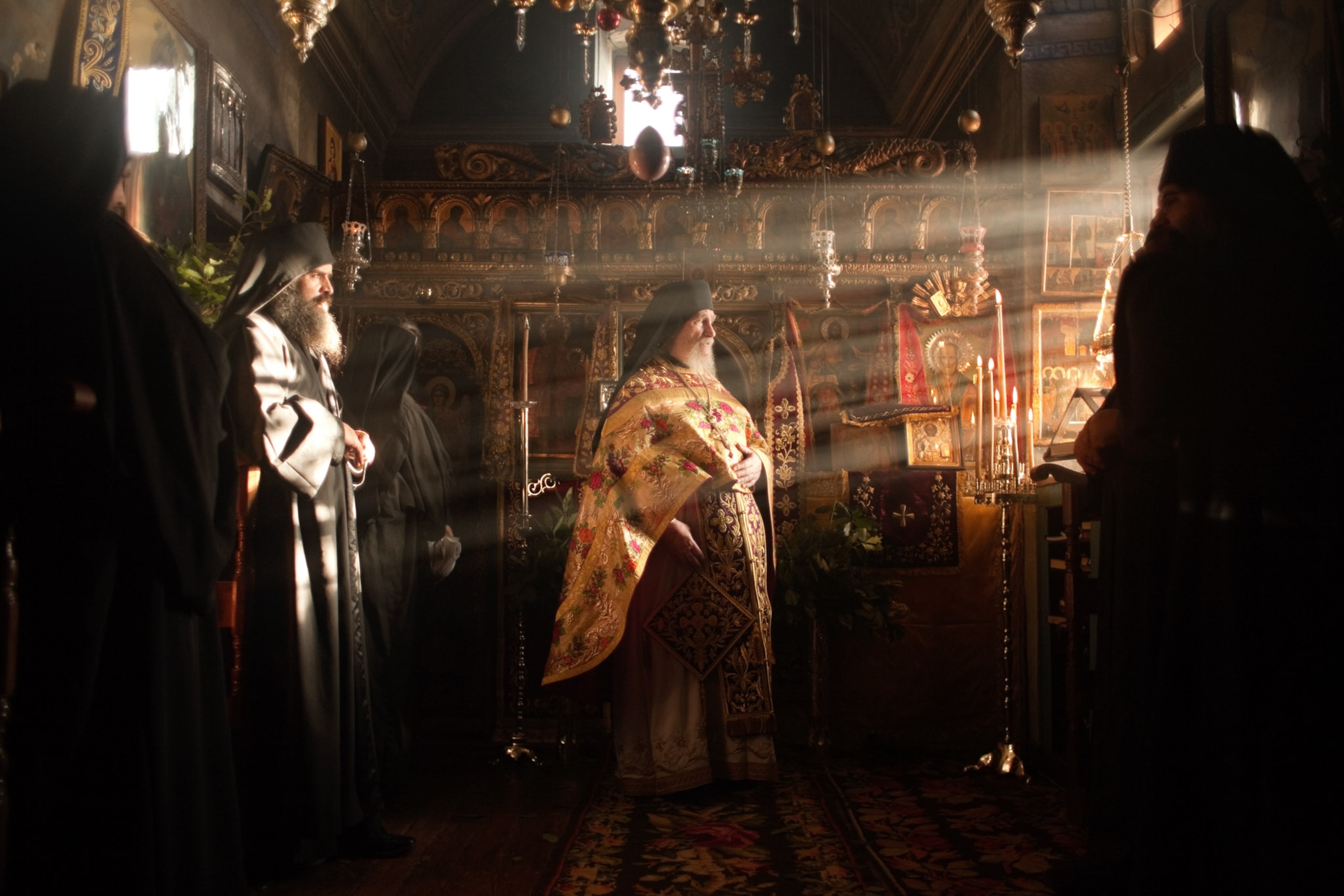 A man in a red gold adorned robe stands center frame as light streams in from the window illuminating him and a mand in a black robe standing behind him.