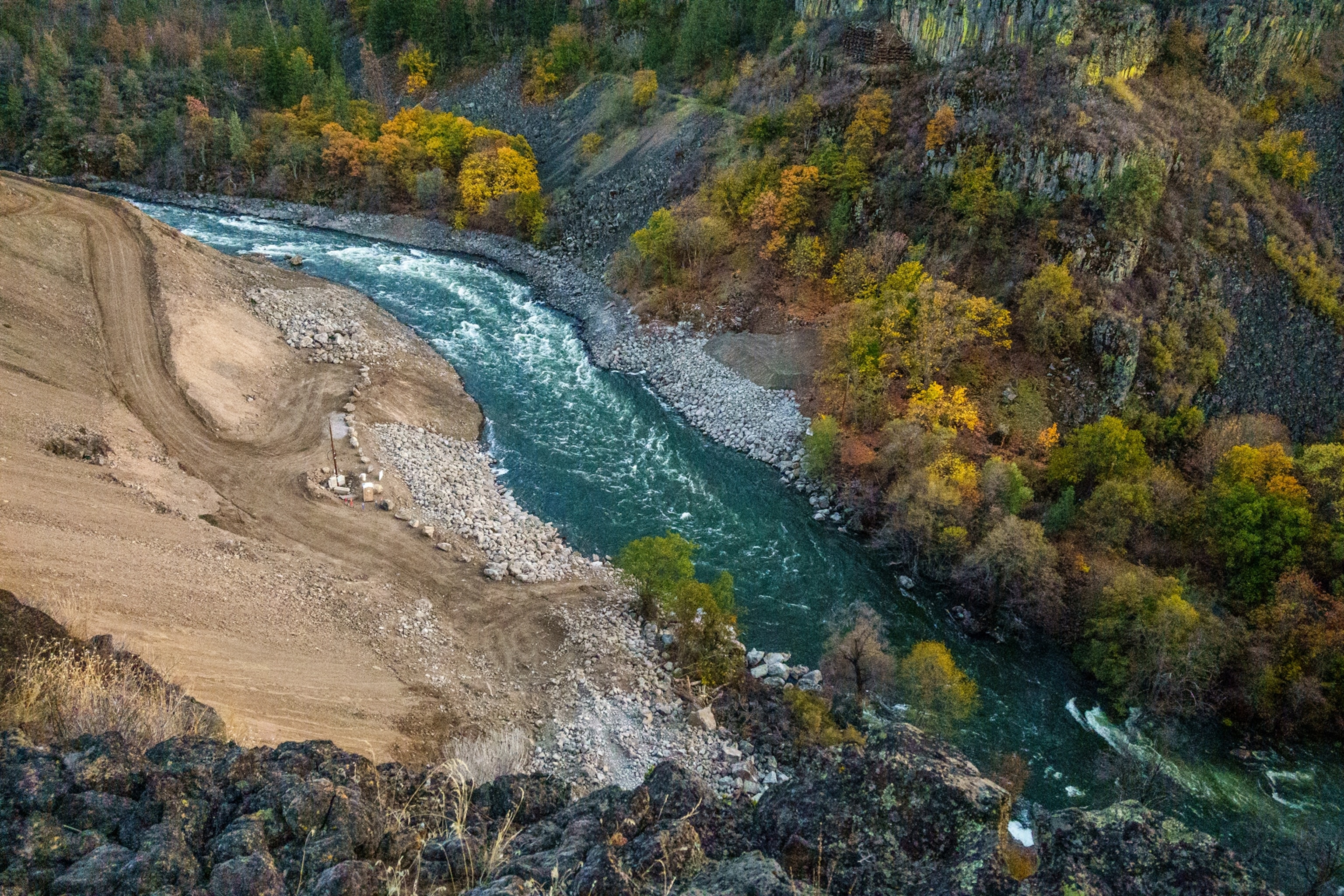 A birds eye view photograph of a river flowing freely. One side is heavily forested, and the other is more barren with dirt and rocks.