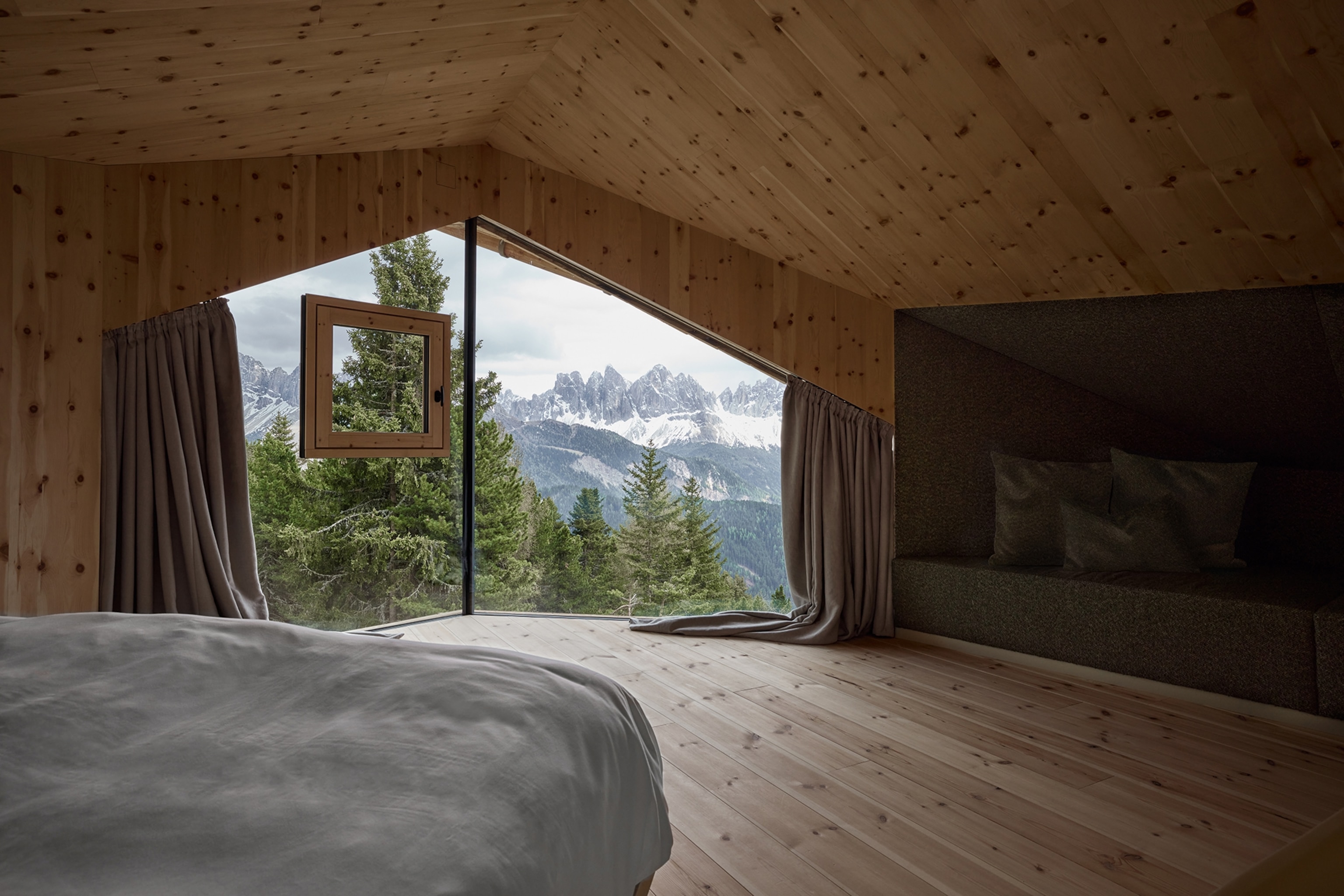Window overlooking snow-capped mountains and trees from a bedroom with wooden walls and flooring