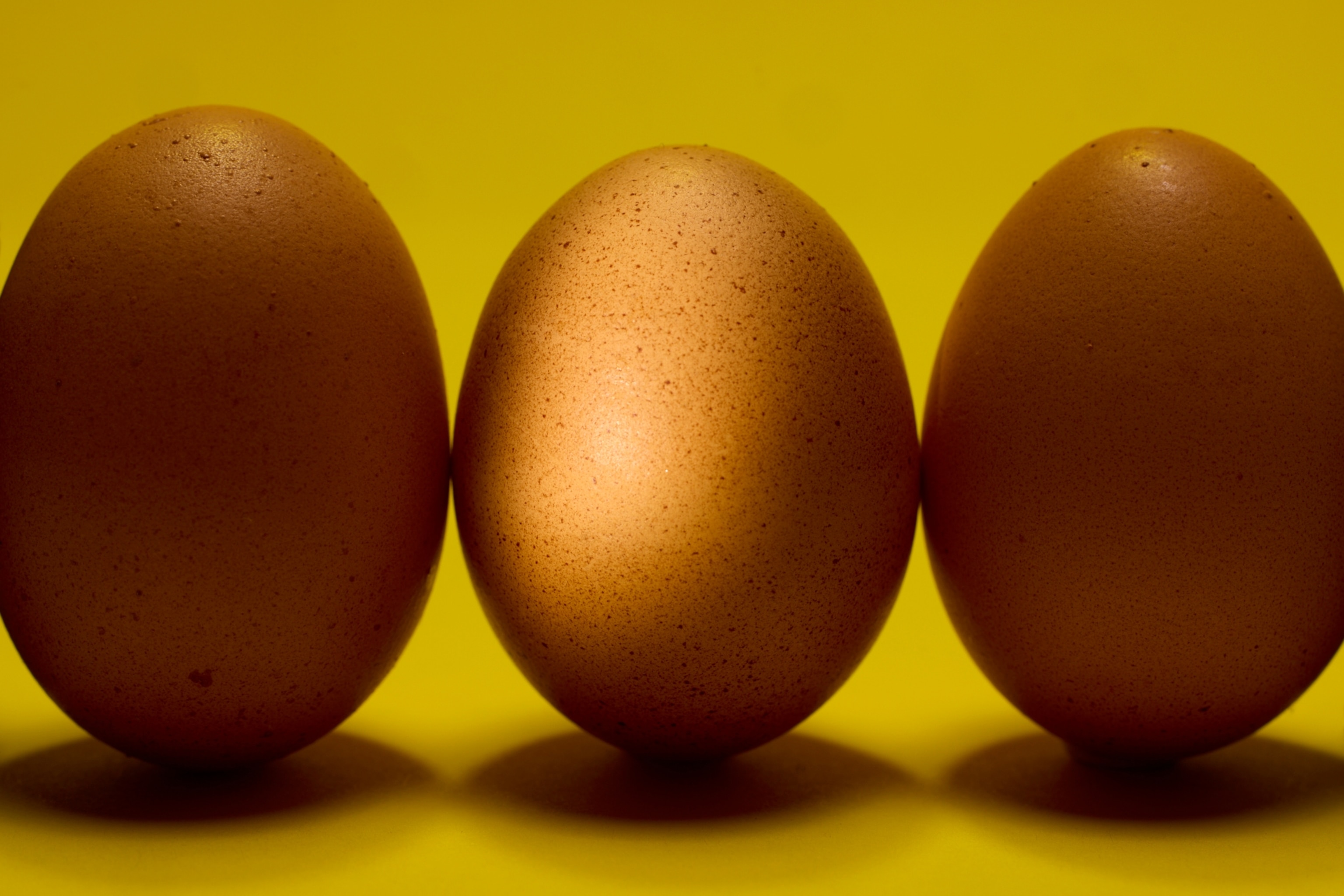 Three brown chicken eggs, the middle one in spotlight, in a line against a yellow backdrop.