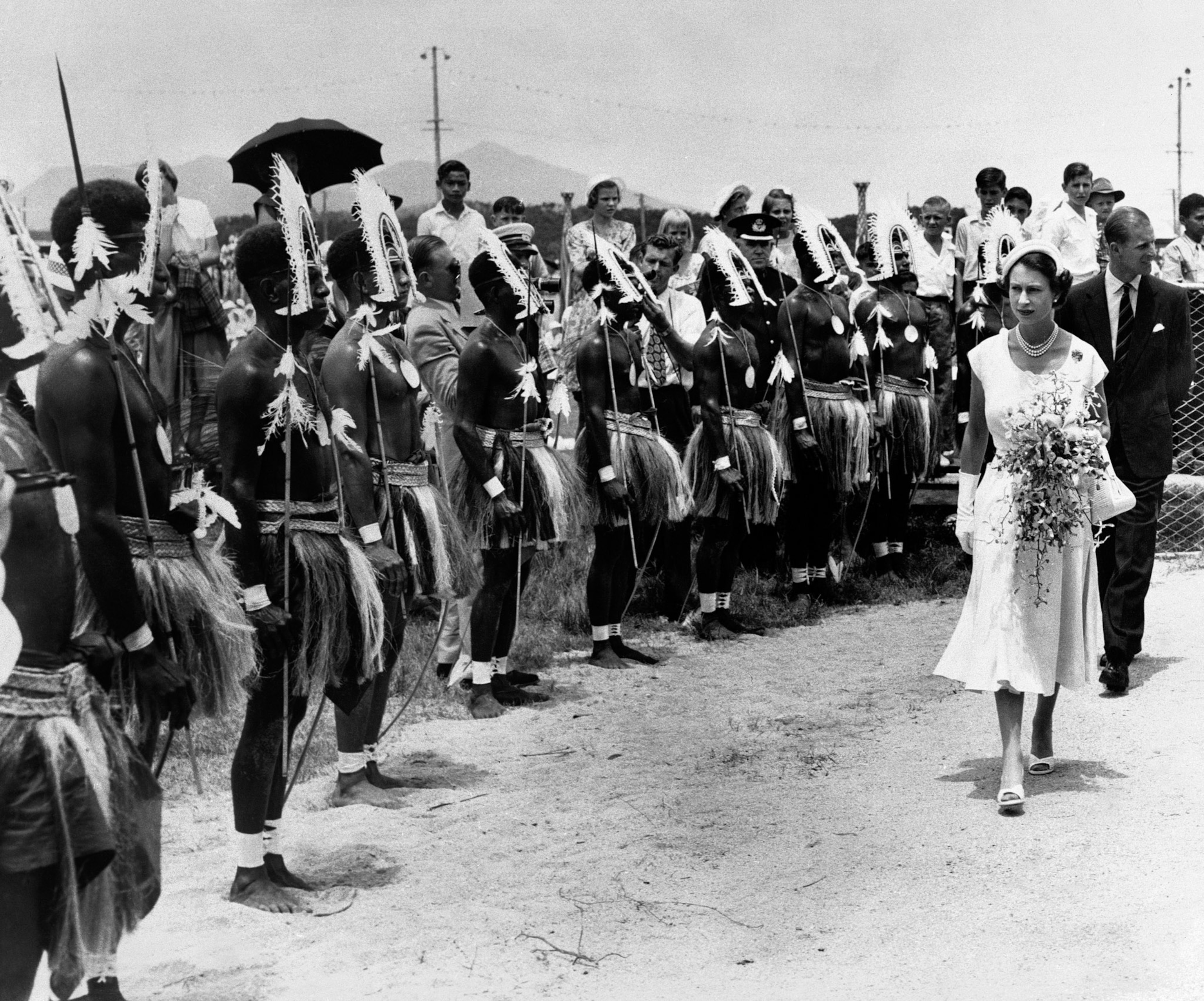 A black and white image that shows a group of people lined up in headdress as the late queen of England looks.