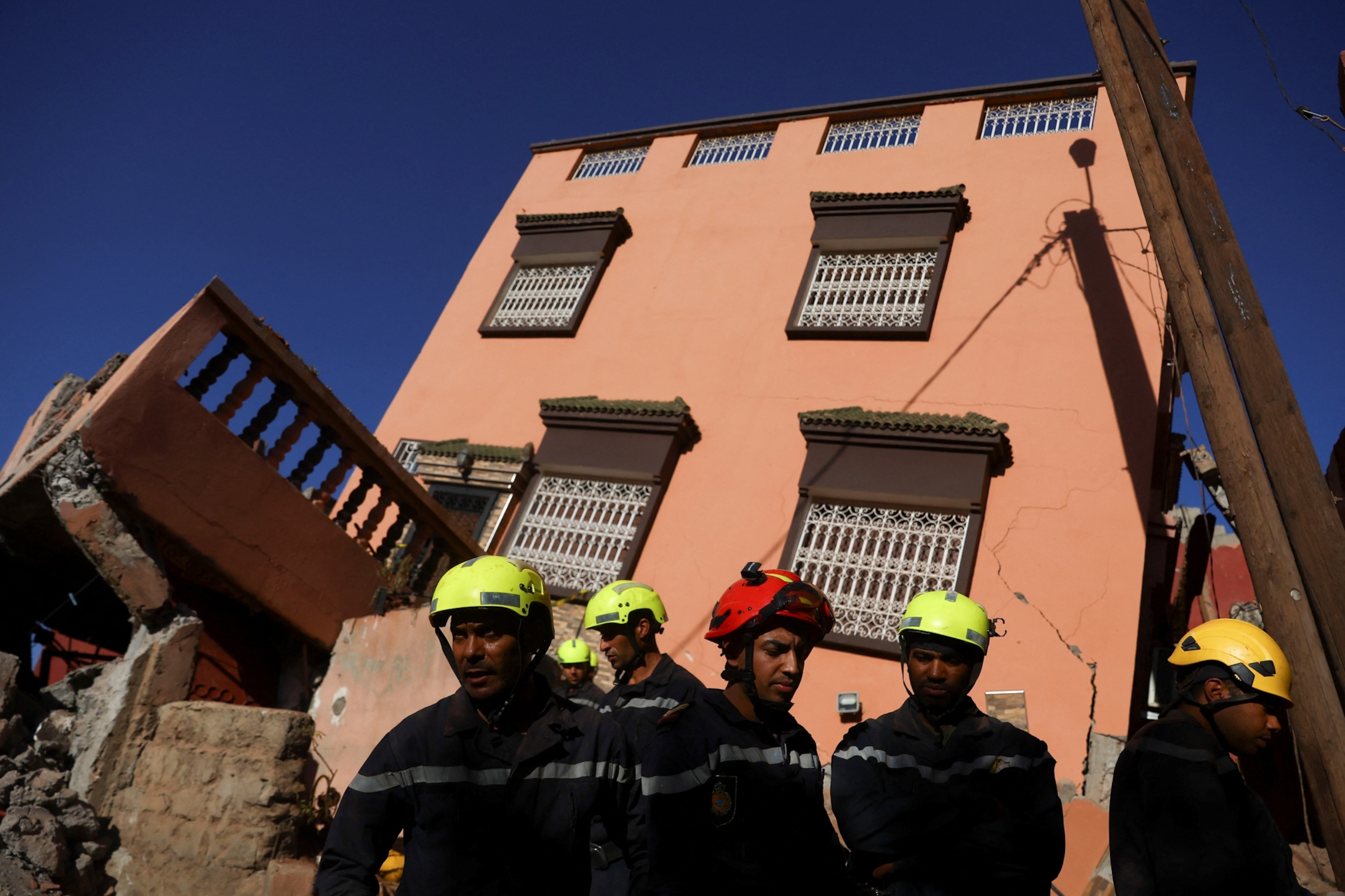 Emergency crew members are seen standing before a peach colored building, which is cracked and fallen off its foundation.
