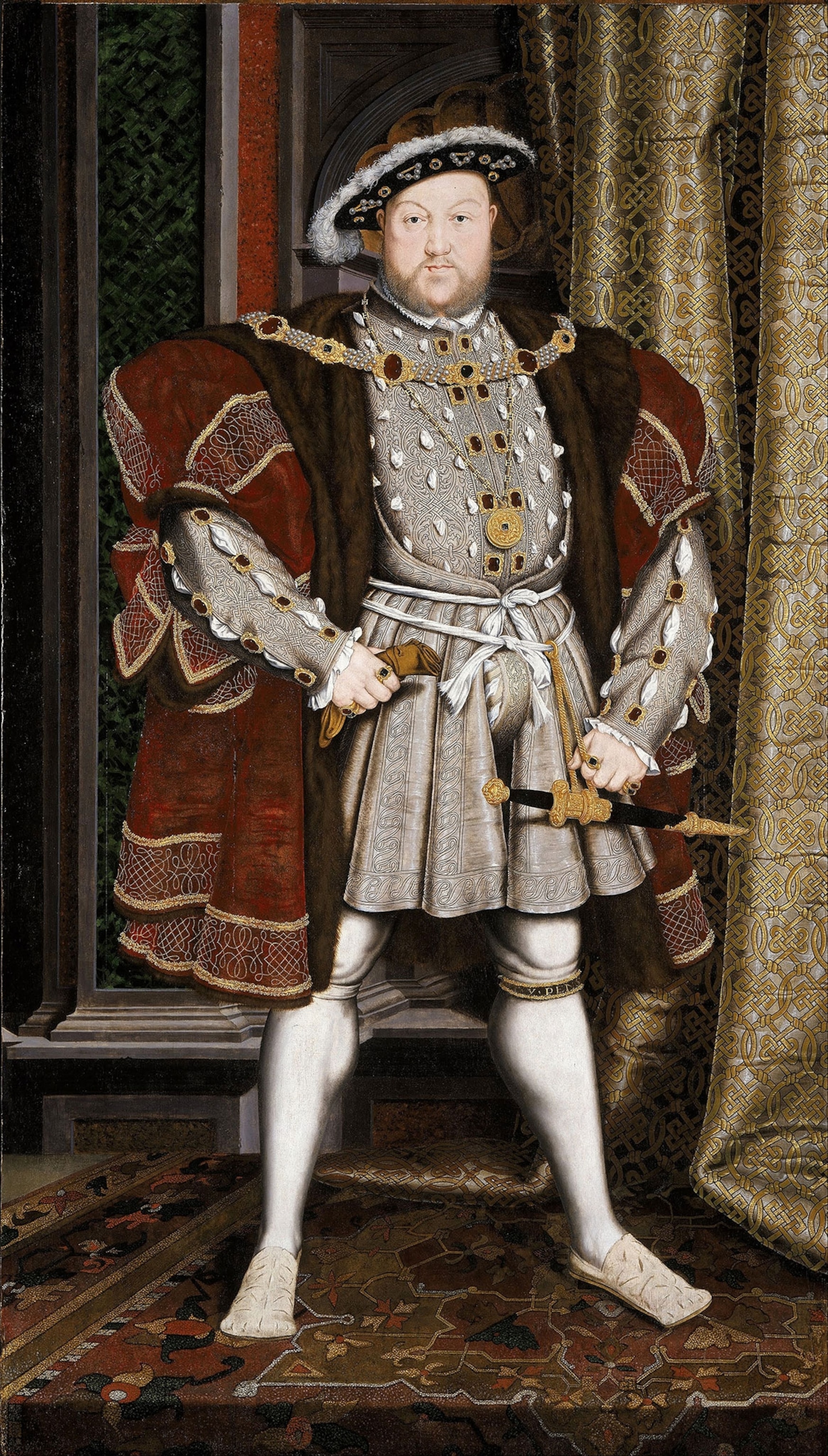 A picture of a man, dressed in an oversized and ornate red coat with wide shoulders and an ornate silver top and dress like bottom with a codpiece. He is also wearing white tights, standing on a multi-colored oriental rug, with gold curtains hanging in the background on the right.