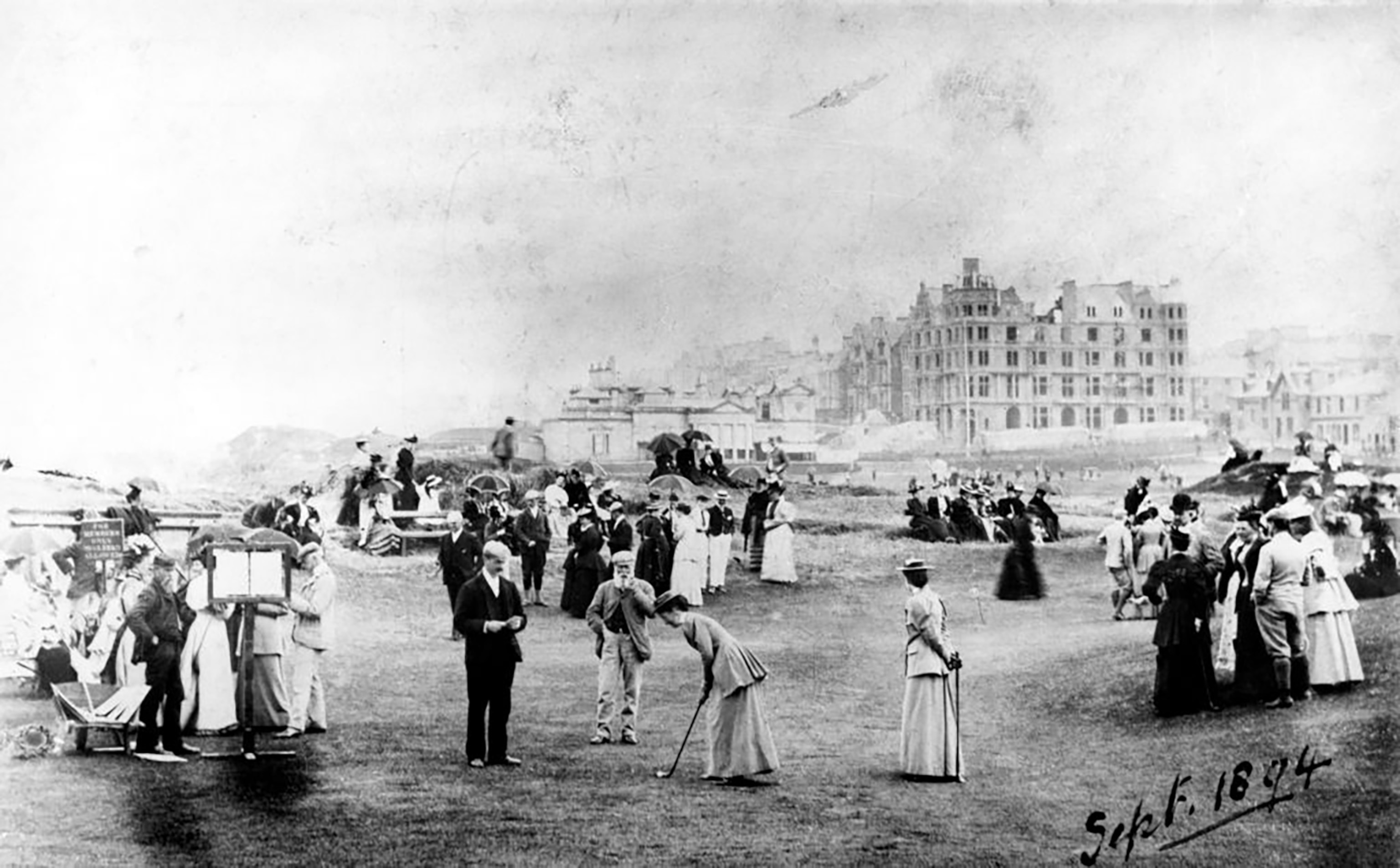 Groups of men and women standing on hummocky ground with buildings in far distance; two women with clubs putting.