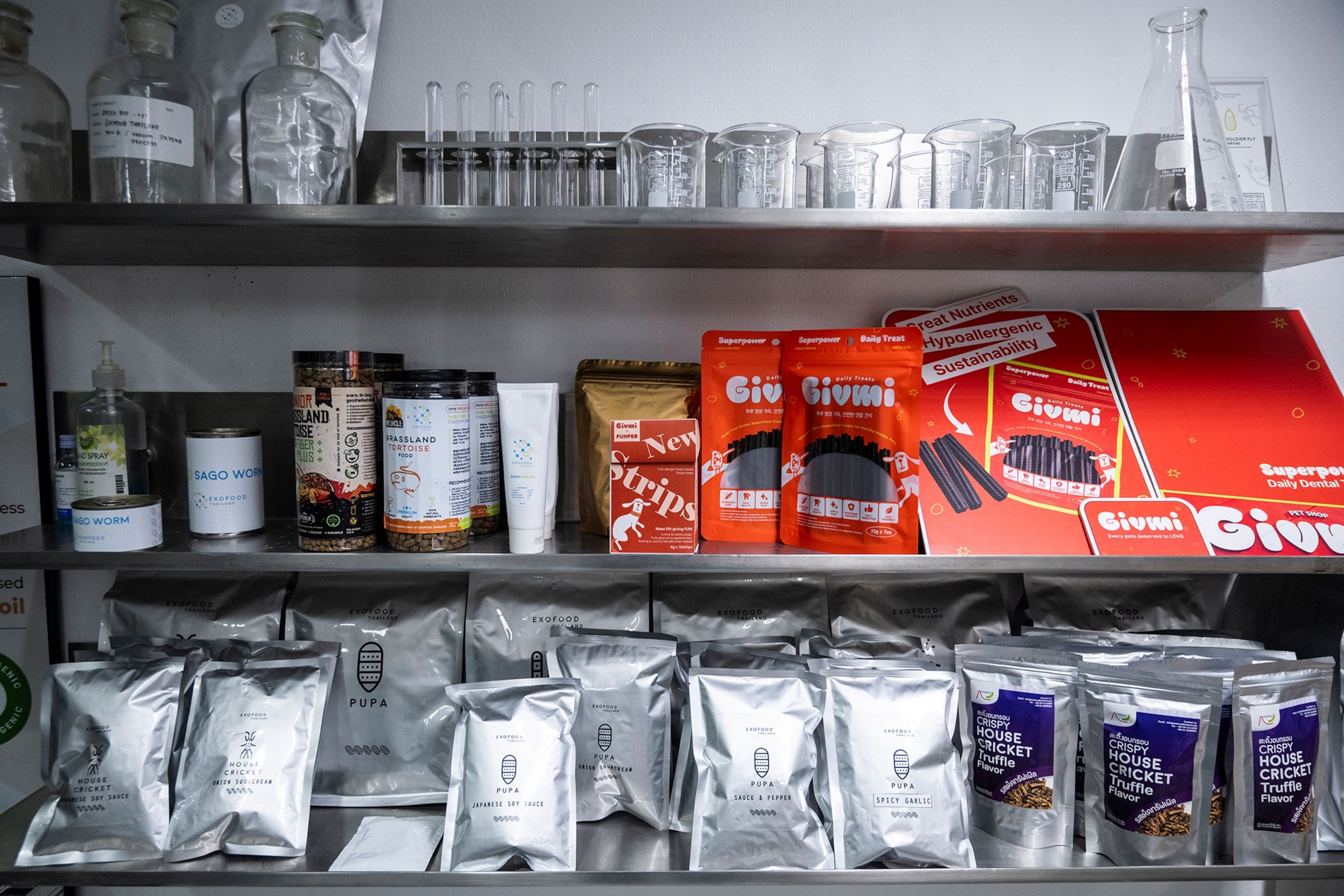 Stacked metal shelves filled with red and silver packages on the bottom two shelves while beakers and flasks fill the top shelf.