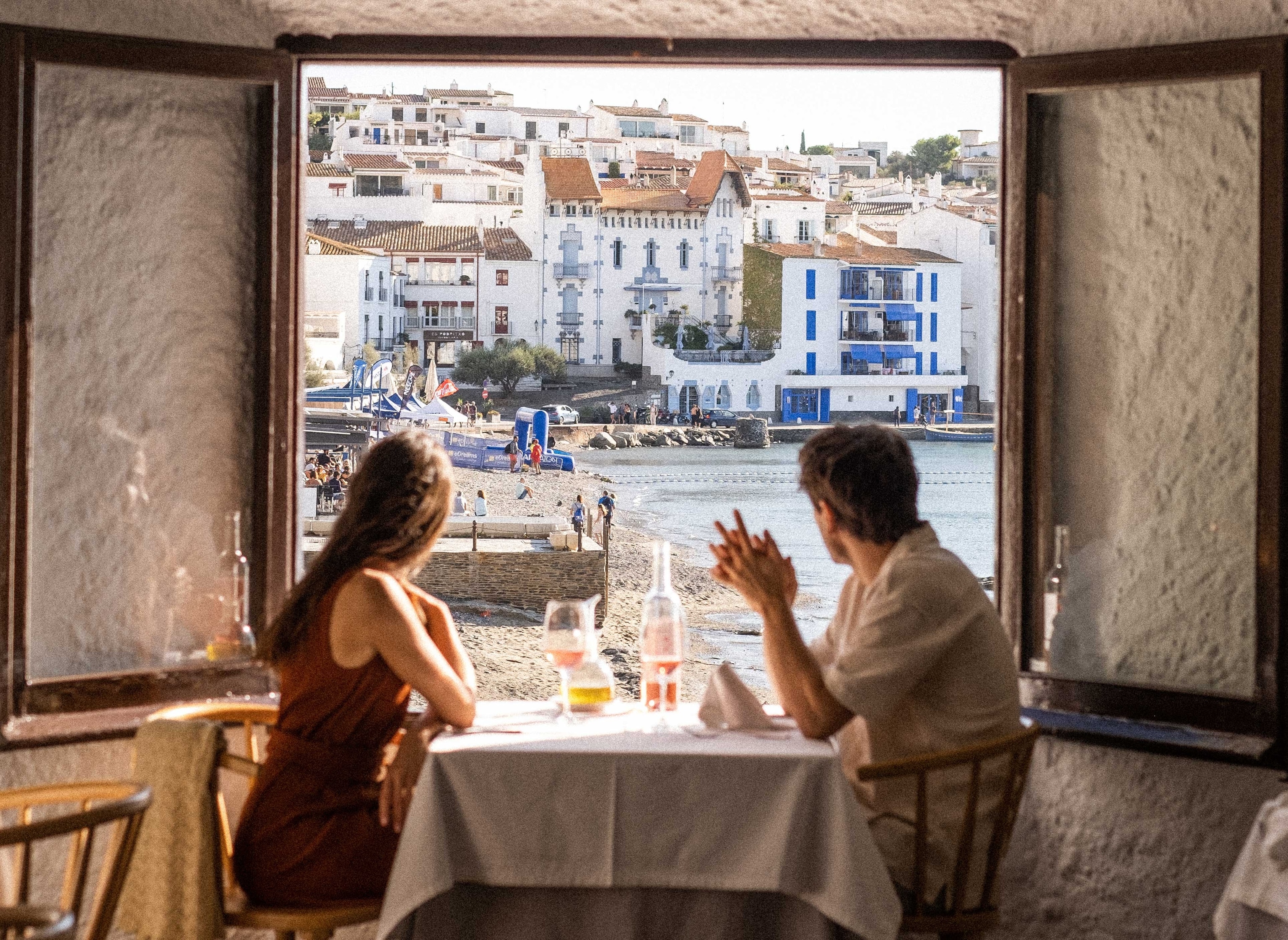 a couple sat at a restaurant table next to a window, overlooking a pretty coastal town and beach