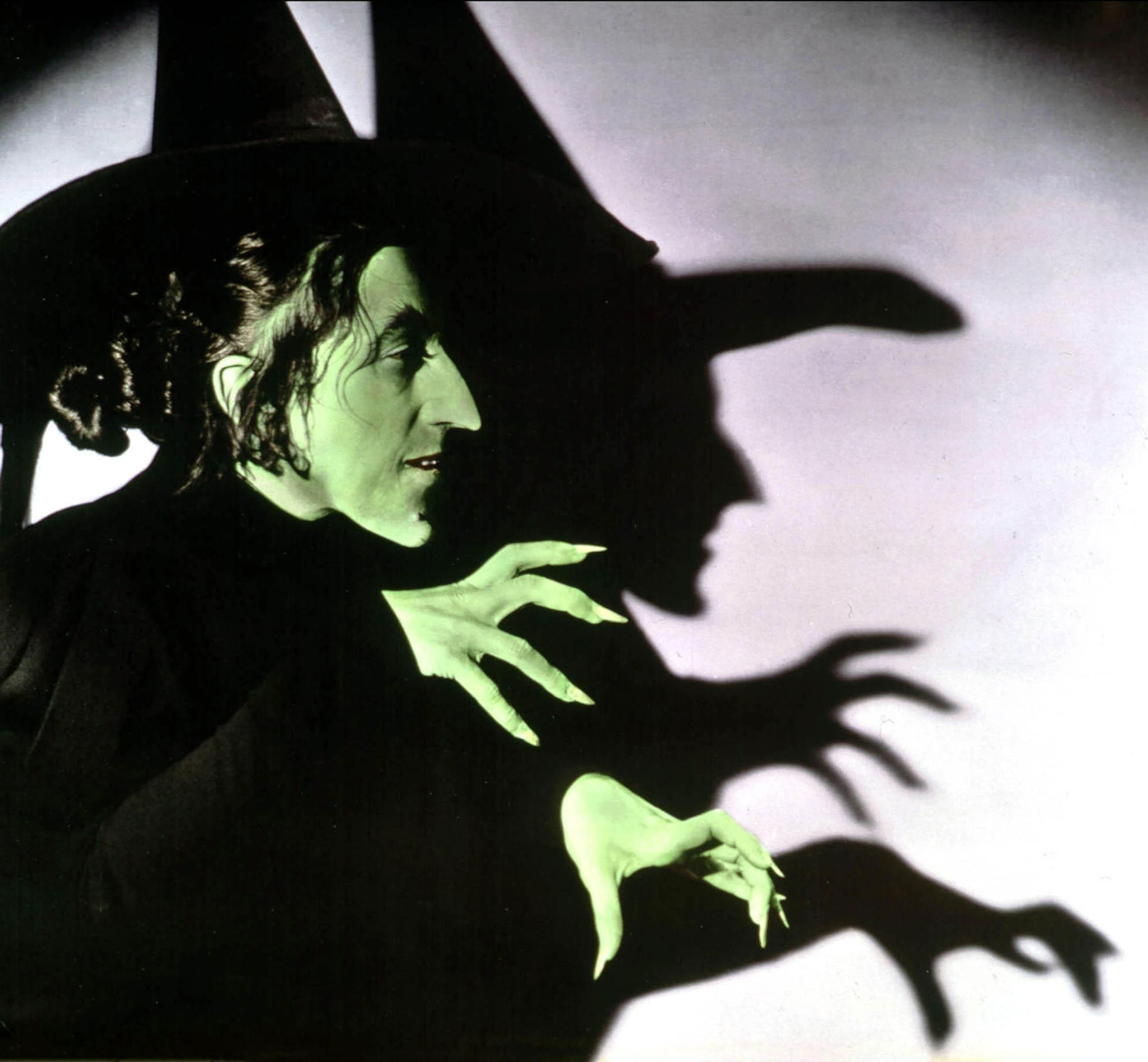 Photo of Wizard of Oz's Wicked Witch of the West with a shadow behind emphasizing her pointy black hat, long finger nails and pointy nose and chin.