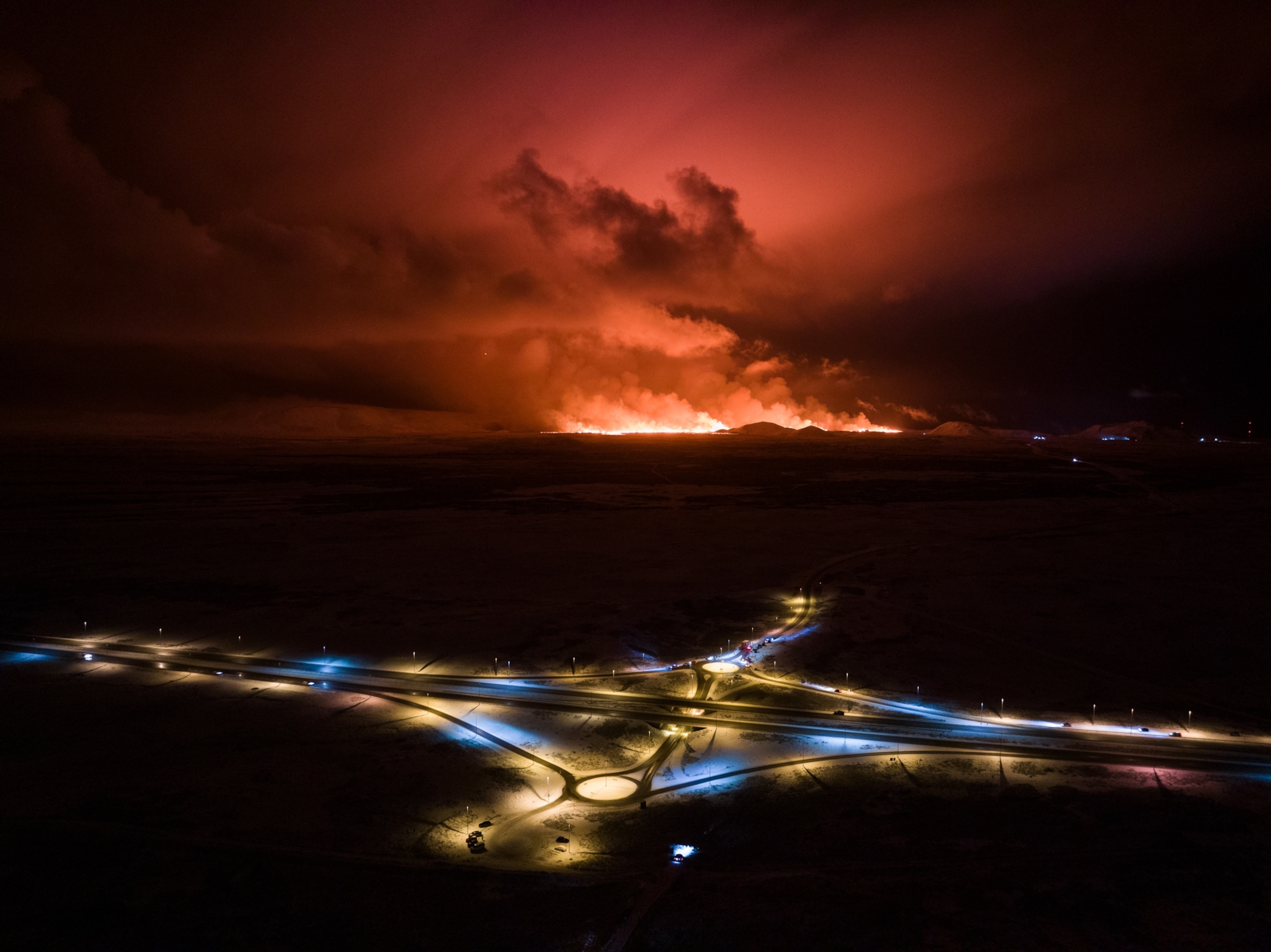 Drone aerial of a road lit by white and blue street lamps with the red glow of a volcanic eruption in the distance.