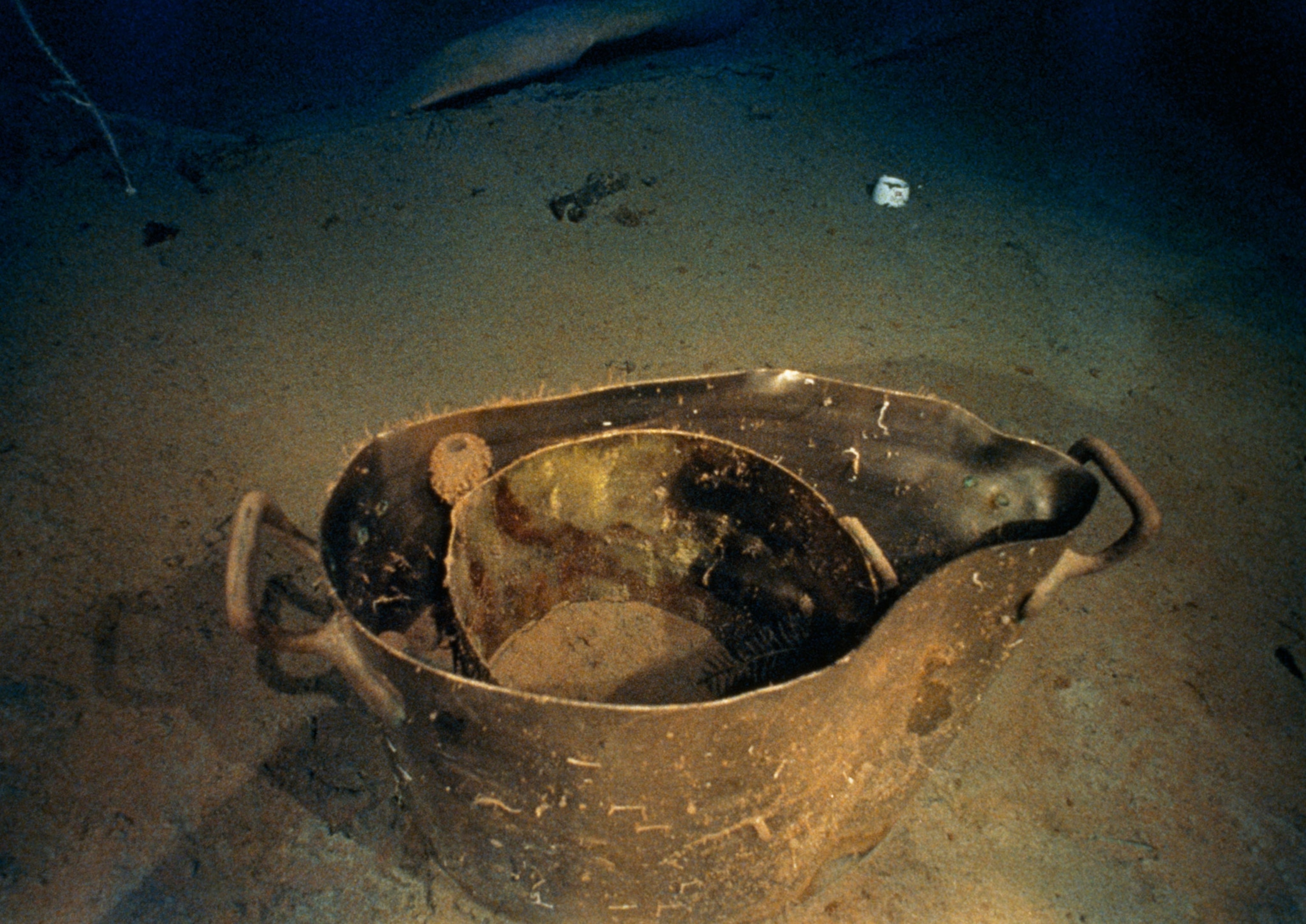 A metal cooking pot with a smaller pot sitting inside, sitting on the ocean floor. Another small white mug is sitting on the floor in the background.