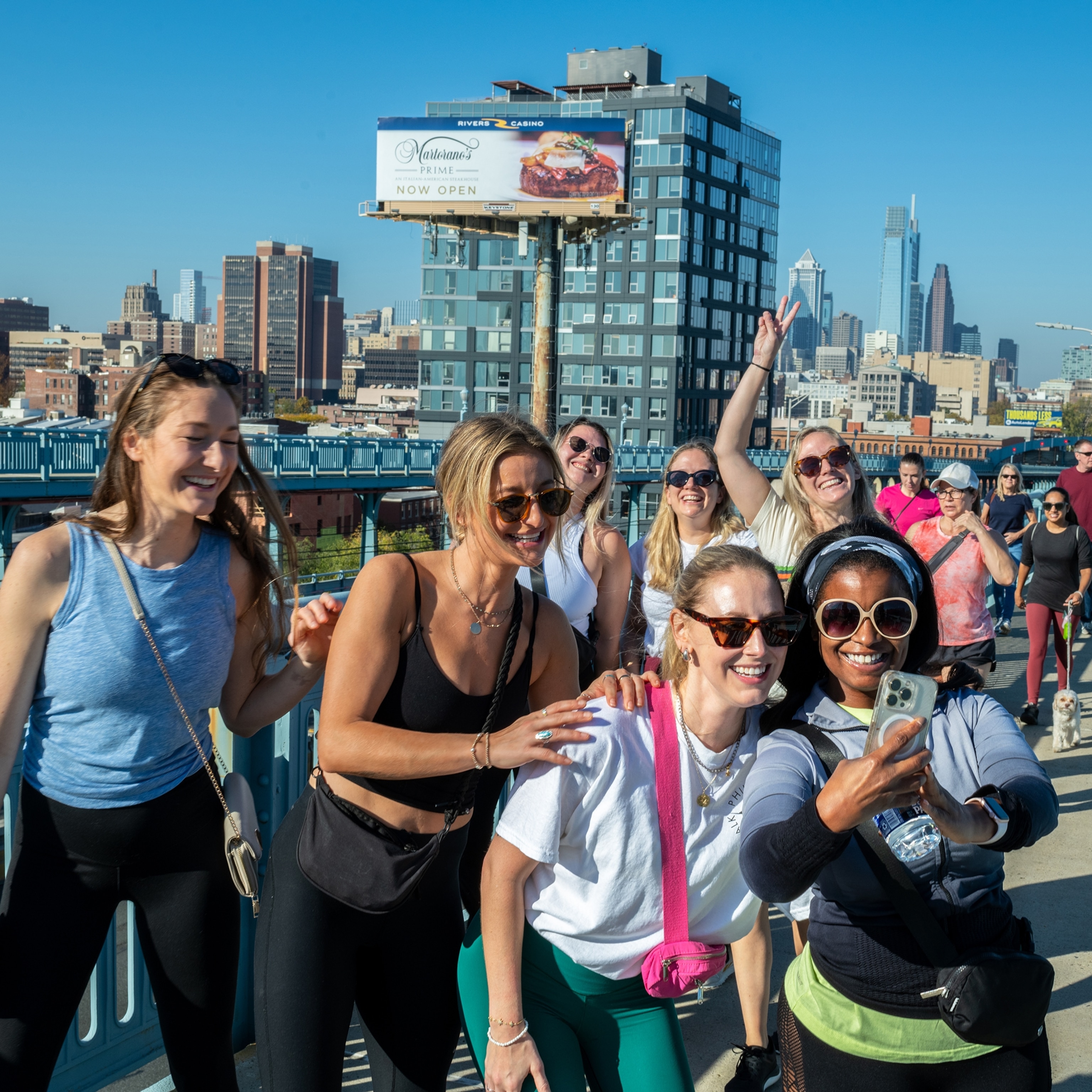 A crowd of people walk over a city bridge. Seven women at the front pose for a group selfie together.