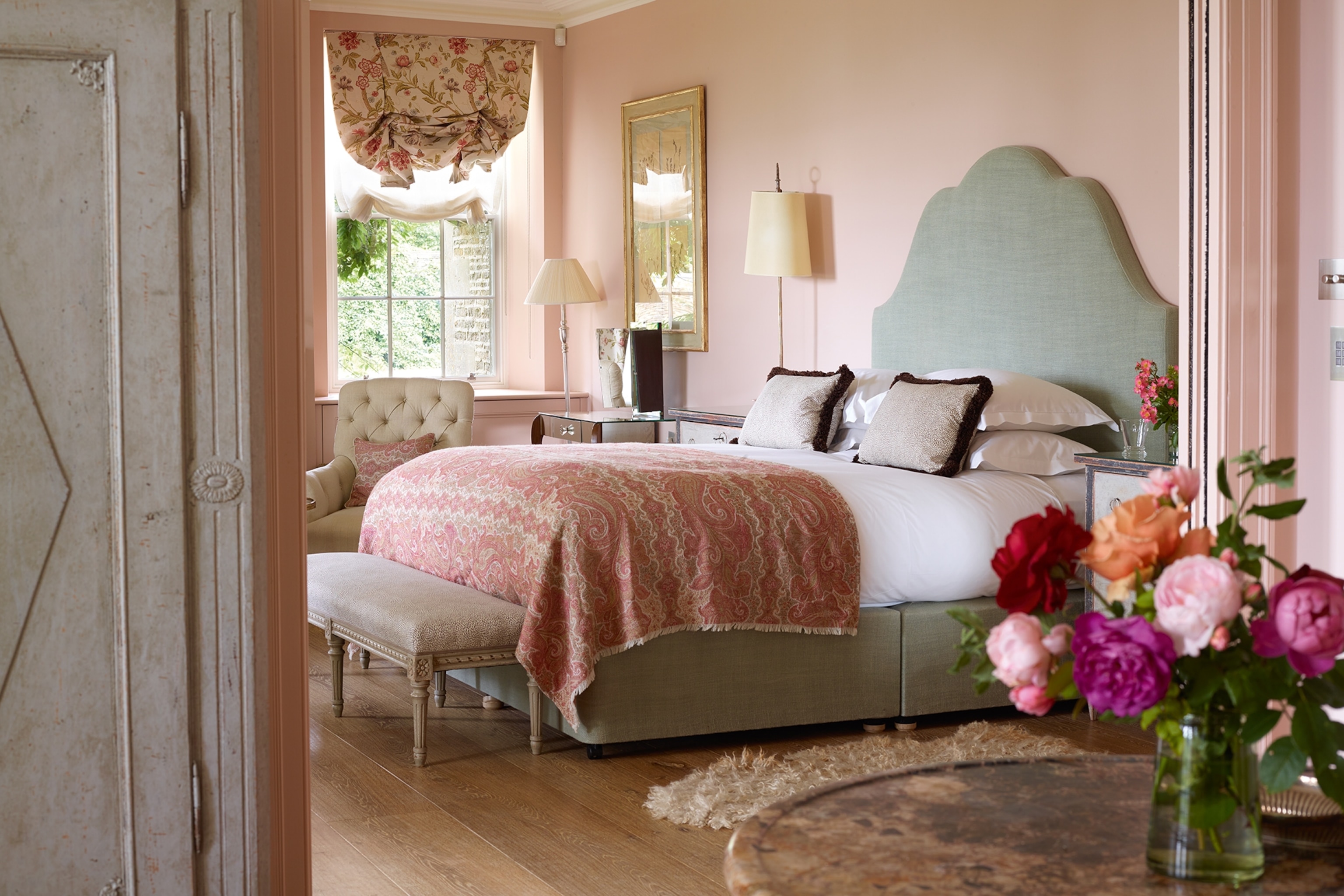 A pink-forward and English rose-themed hotel bedroom with a bunch of fresh peonies in a vase.