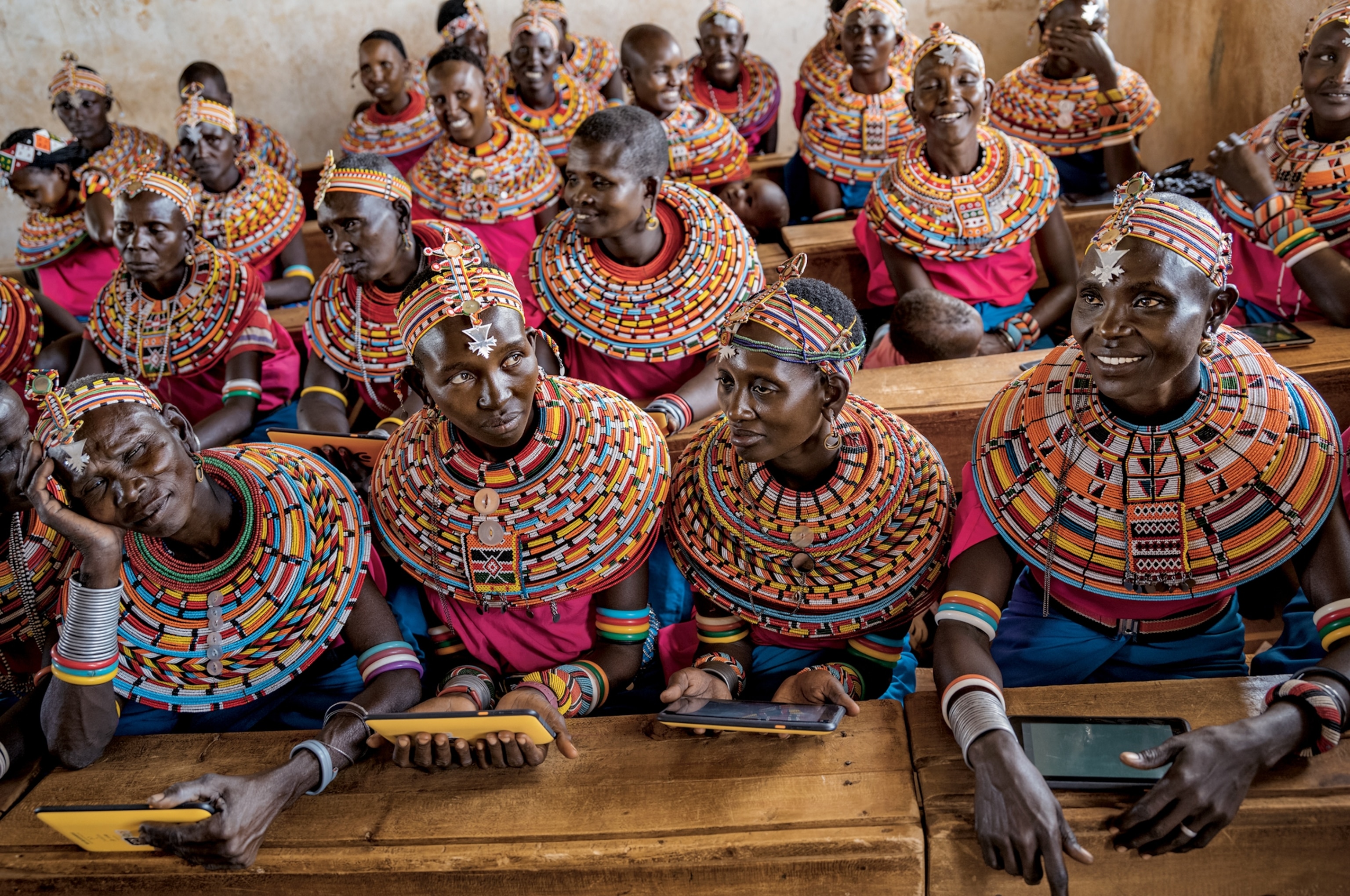 women in colorful dresses sitting in classroom and holding tablets.