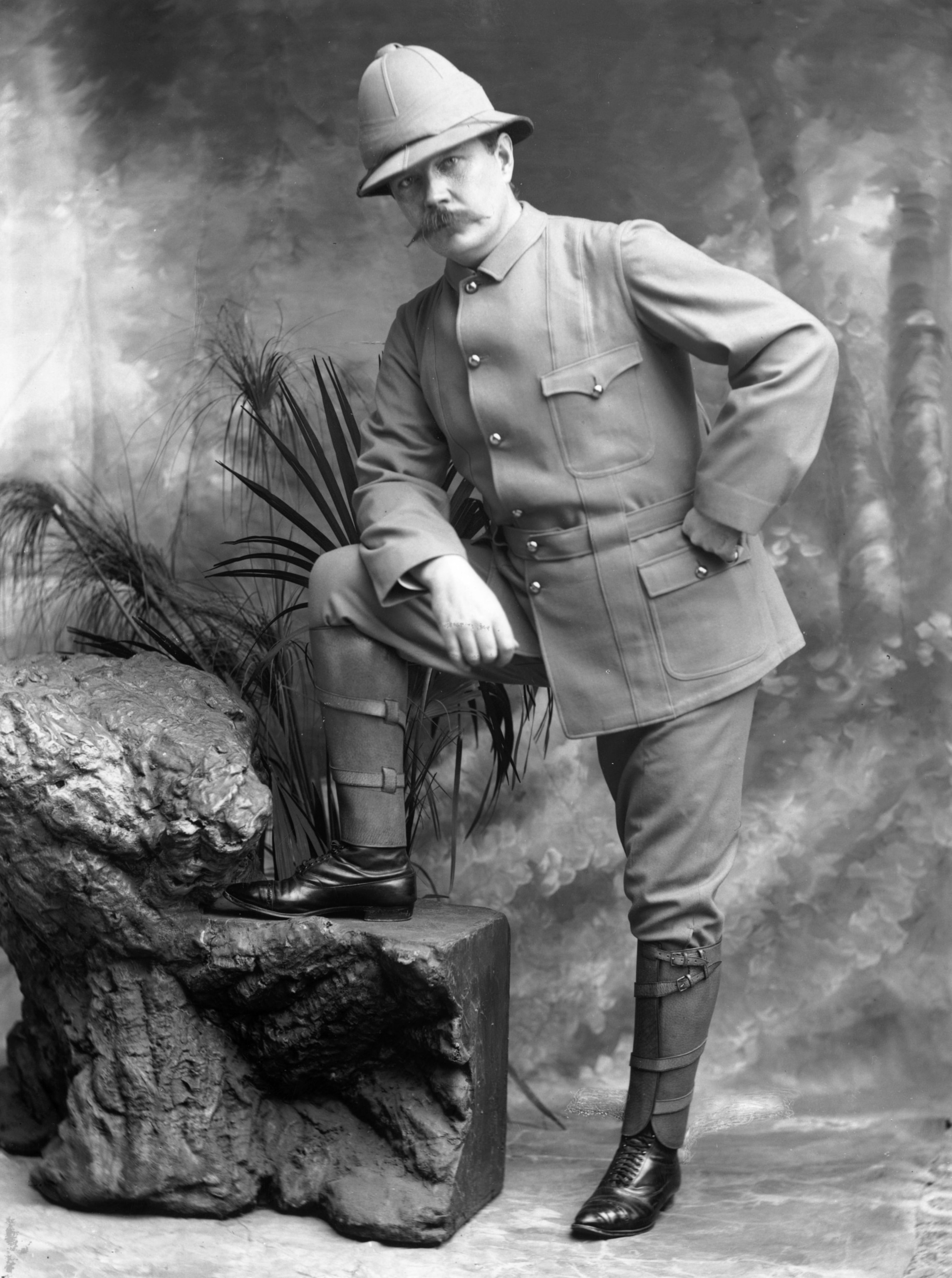 A mustachioed white man wearing a 19th-century British military uniform poses in front of a photo studio backdrop with one foot on top a large rock.