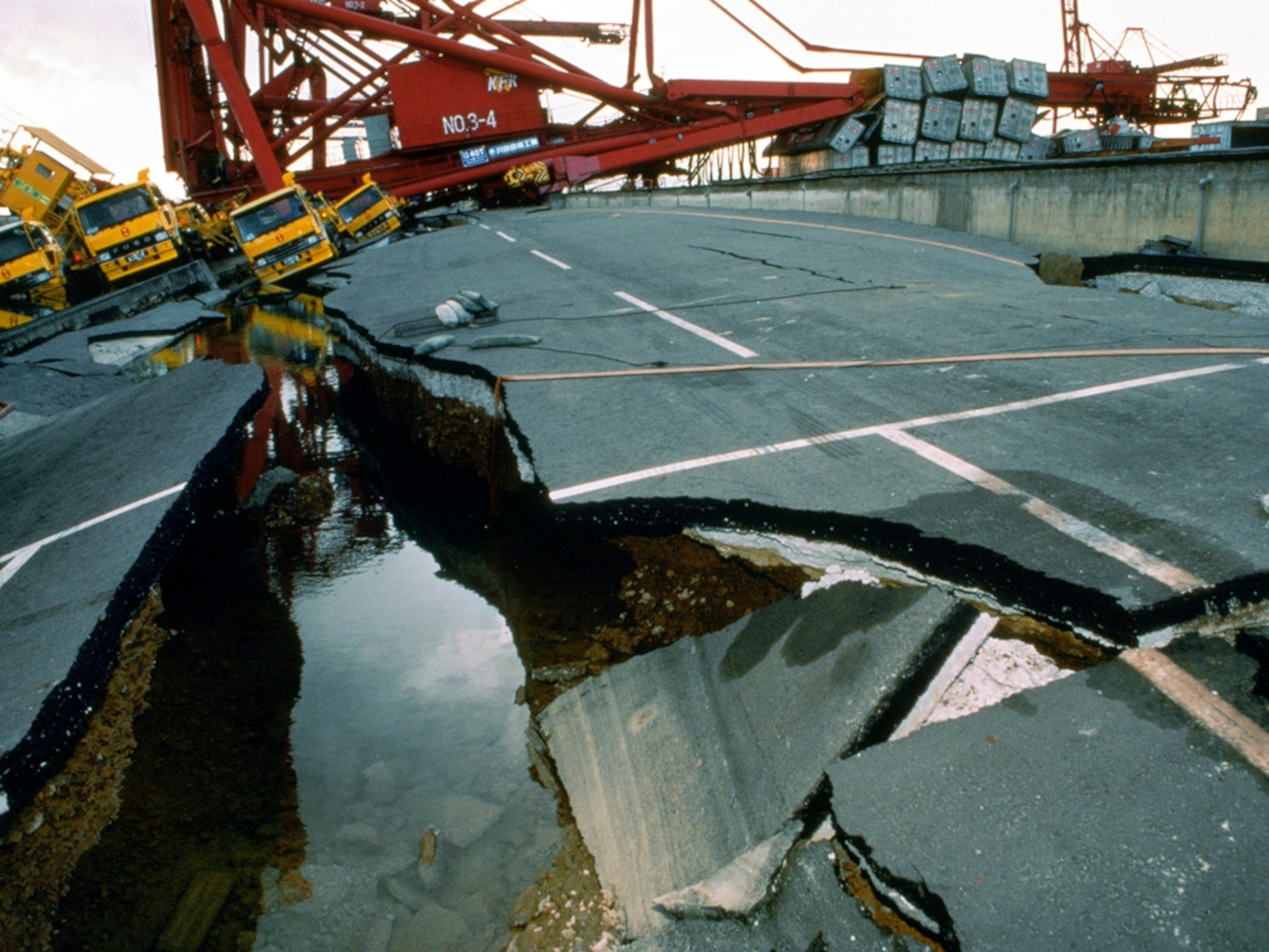 An earthquake has fractured an asphalt road, thrown trucks into a pile, and toppled a giant crane.