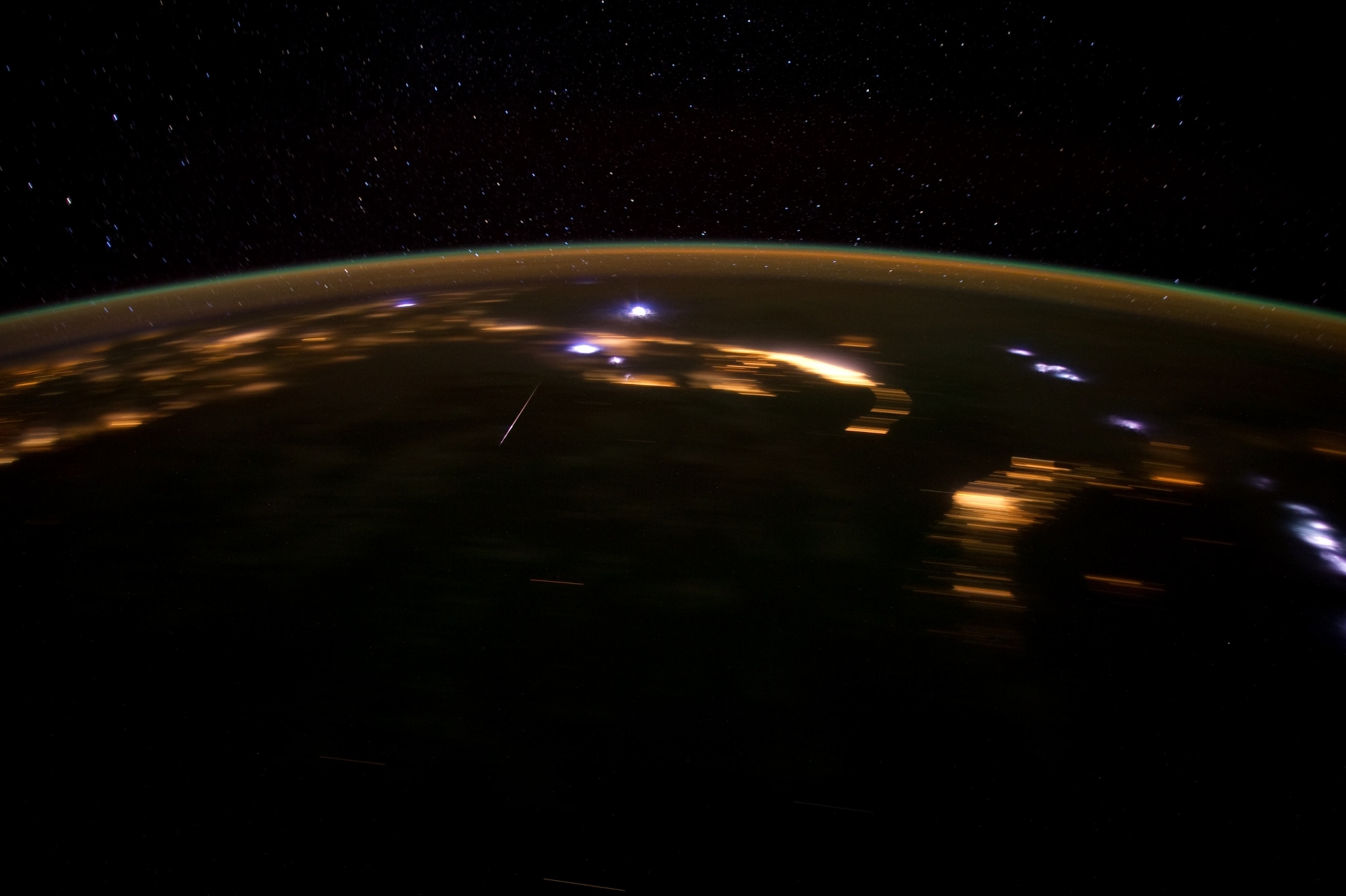 A view from the ISS looking out over Earth with its visible atmosphere. A streak of a meteor is seen over a dark patch of Earth.